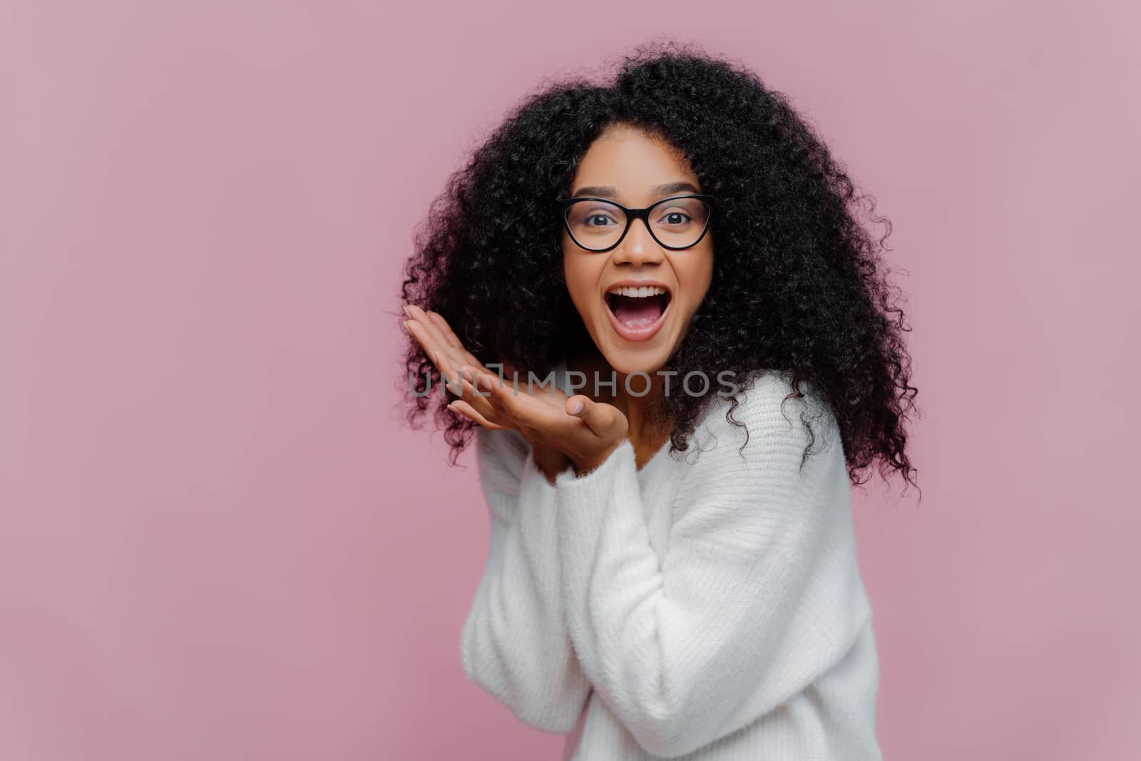 Overjoyed happy curly young woman with cheerful expression, keeps mouth widely opened, wears casual white jumper and optical glasses, poses over violet background. Emotions and happiness concept by vkstock