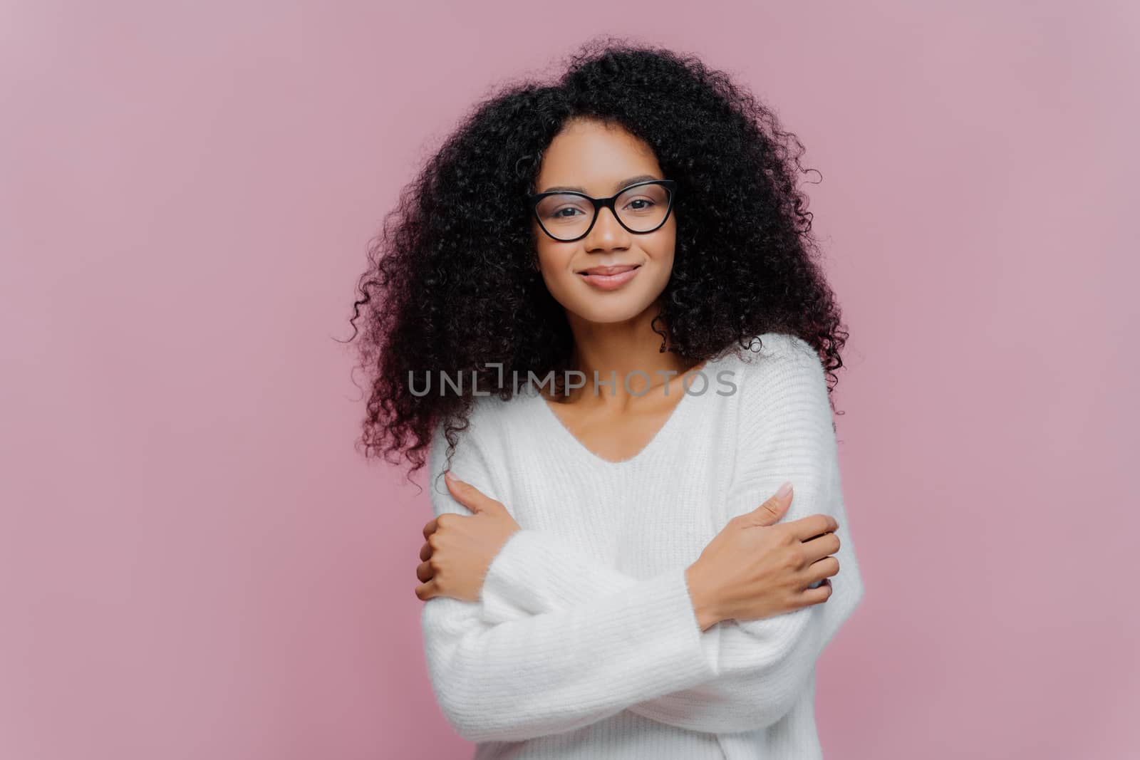 Pleasant looking millennial Afro American woman keeps hands crossed over chest, has curly bushy hair, wears white comfortable sweater and spectacles, isolated over purple background, feels pleased