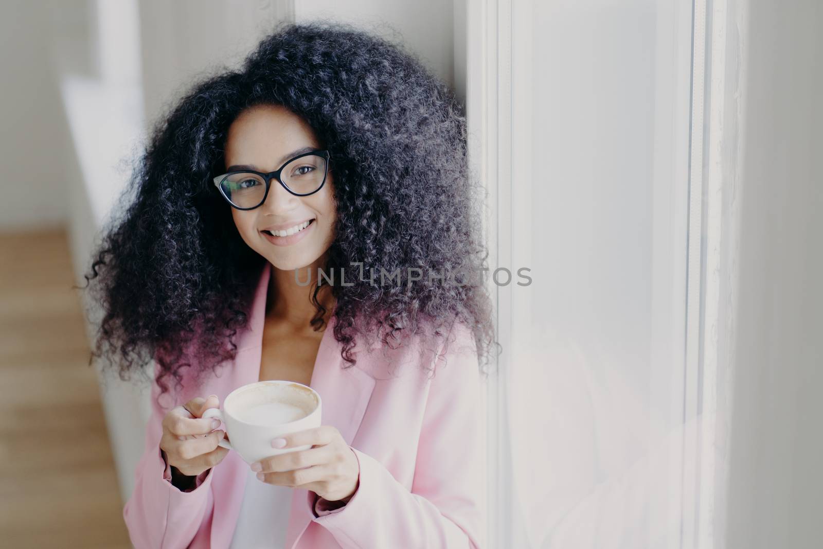 Photo of pleasant looking curly haired lady with toothy smile, dressed formally, poses indoor, holds mug of latte, has coffee break after long hours office work, looks through eyewear, feels happy by vkstock