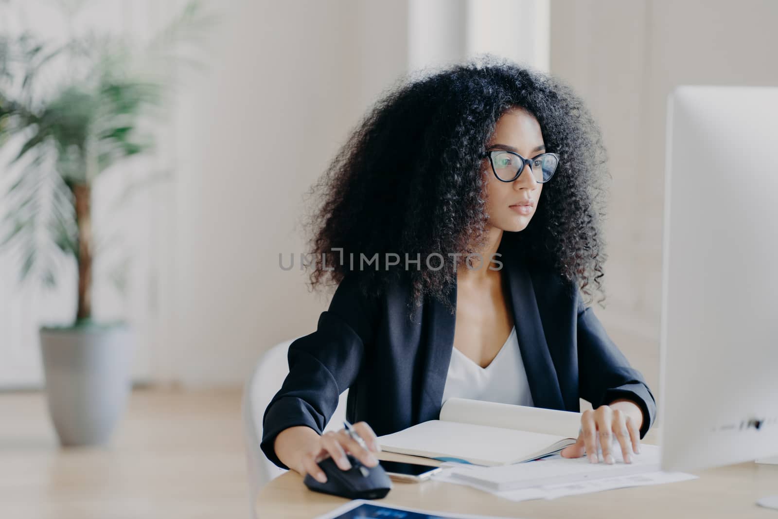 Serious curly businesswoman focused at display of computer, works on making project, surrounded with textbook and papers, wears glasses for vision correction, black suit, poses in office room