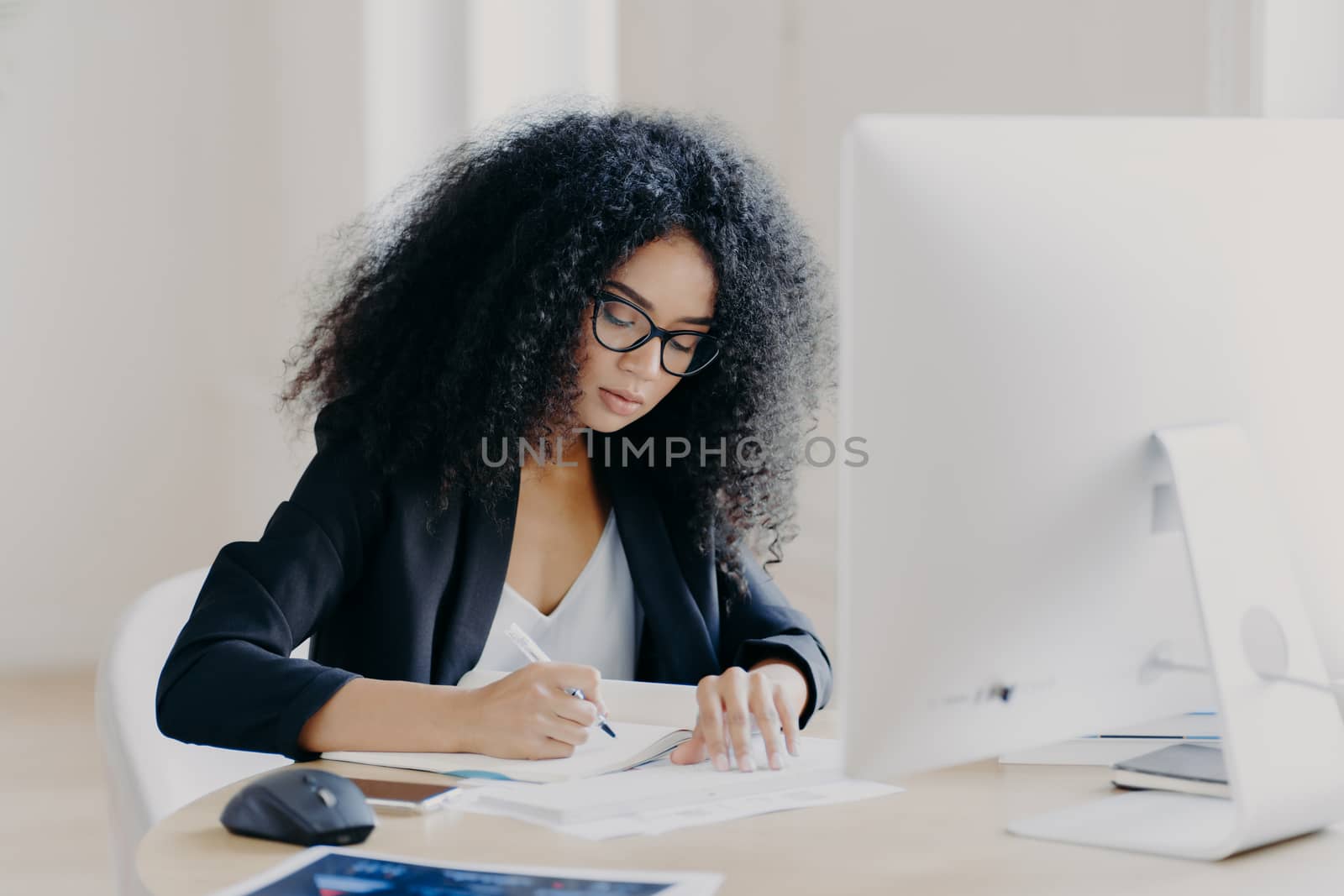 Photo of serious Afro woman writes in papers, sits at table with modern computer, creats article in newspaper, wears glasses and eyewear, poses indoor, works remotely. People and job concept by vkstock