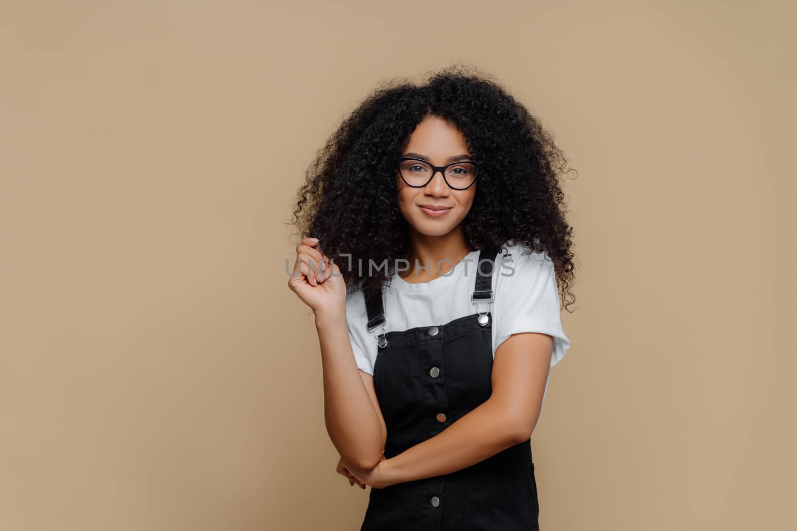 Isolated shot of pleasant looking young African American woman touches curly hair, has bushy hairstyle, wears optical glasses, white t shirt and black overalls, poses over brown studio background by vkstock