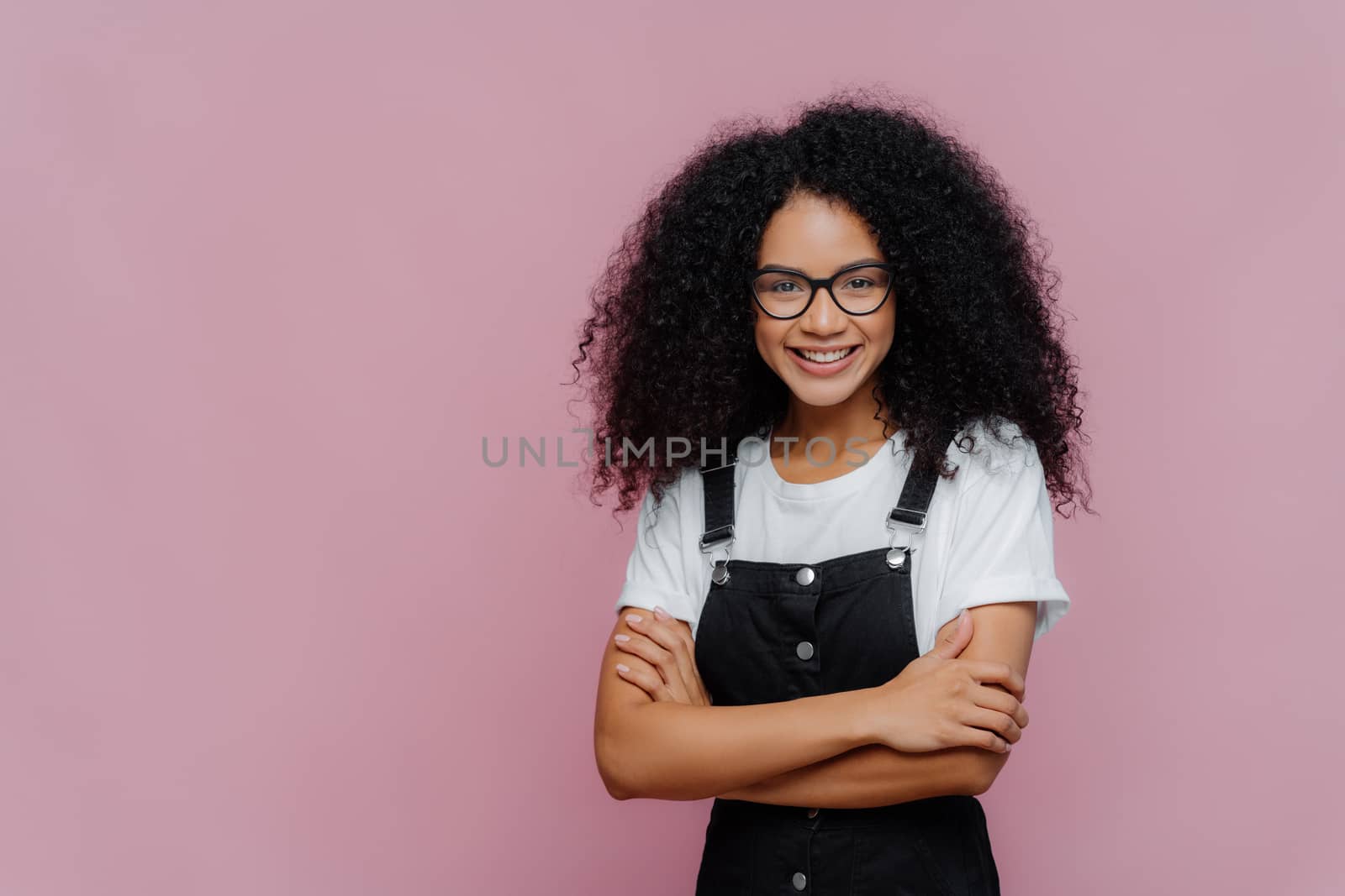 Photo of lovely teenage girl with Afro haircut, keeps arms folded, wears glasses, white t shirt and black overalls, looks gladfully at camera, stands against purple background with copy space for text