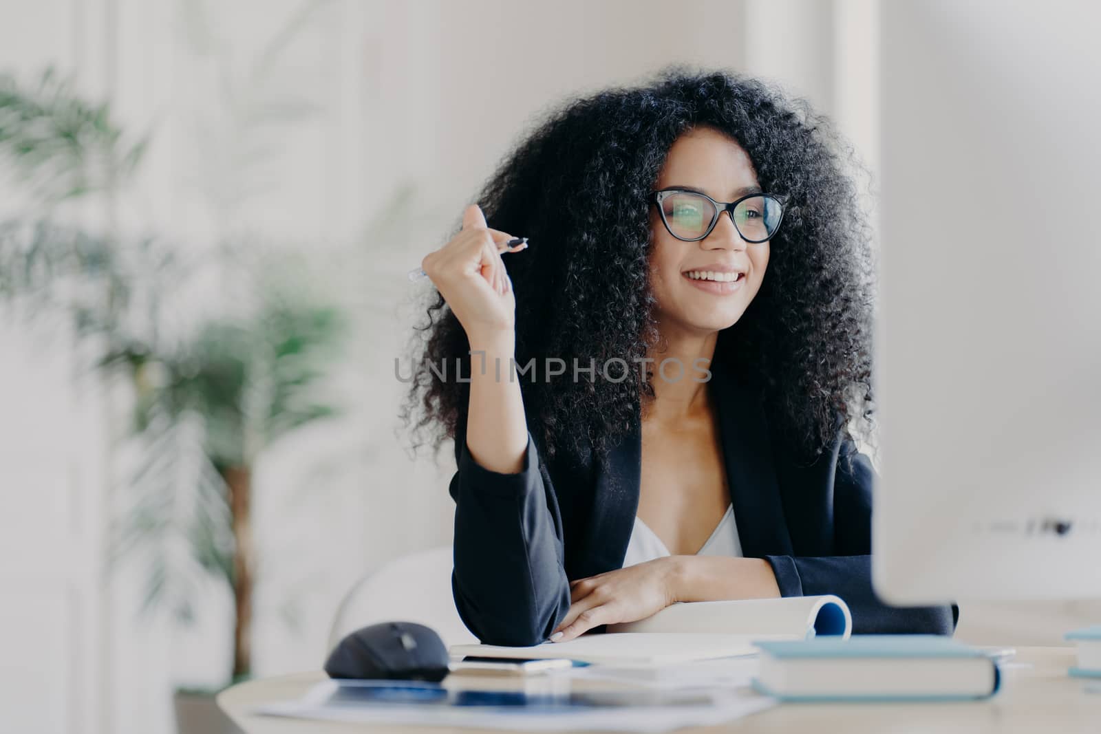 Pretty Afro American woman looks positively at display of computer, writes down information from internet, wears optical glasses and black suit, sits at desktop with necessary things for work