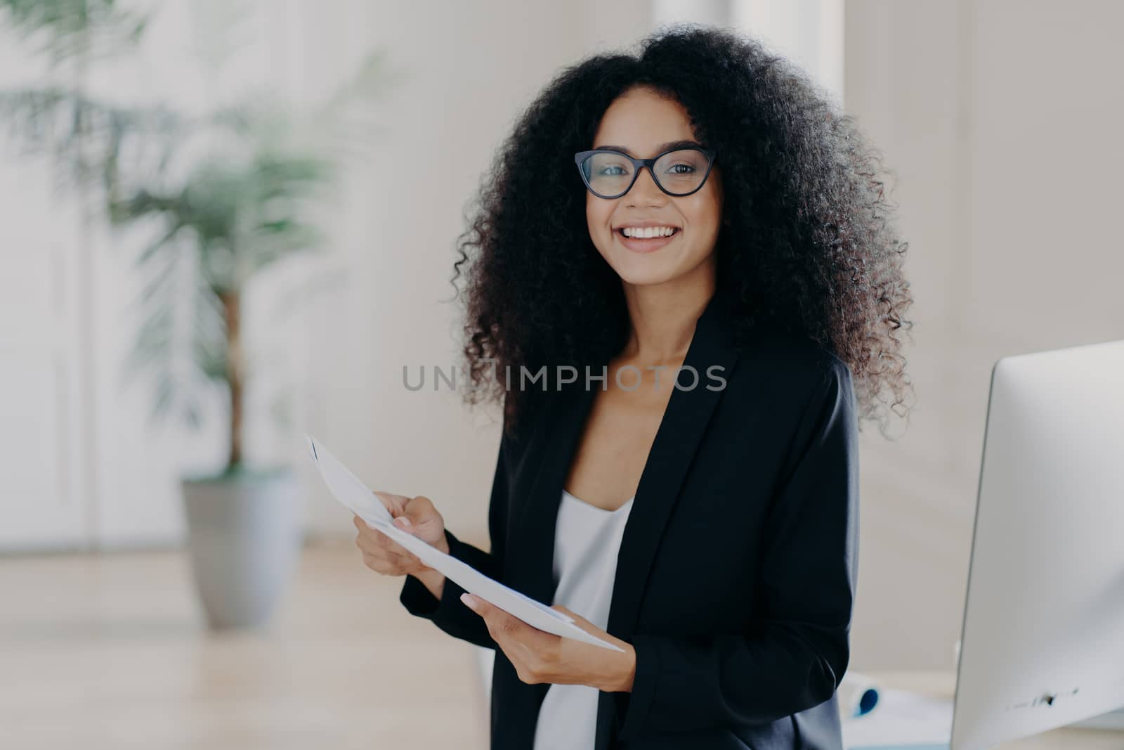 Photo of happy female entrepreneur with Afro hairstyle, studies documentation, wears spectacles and elegant clothes, stands in office interior, prepares to present her business ideas for colleagues by vkstock