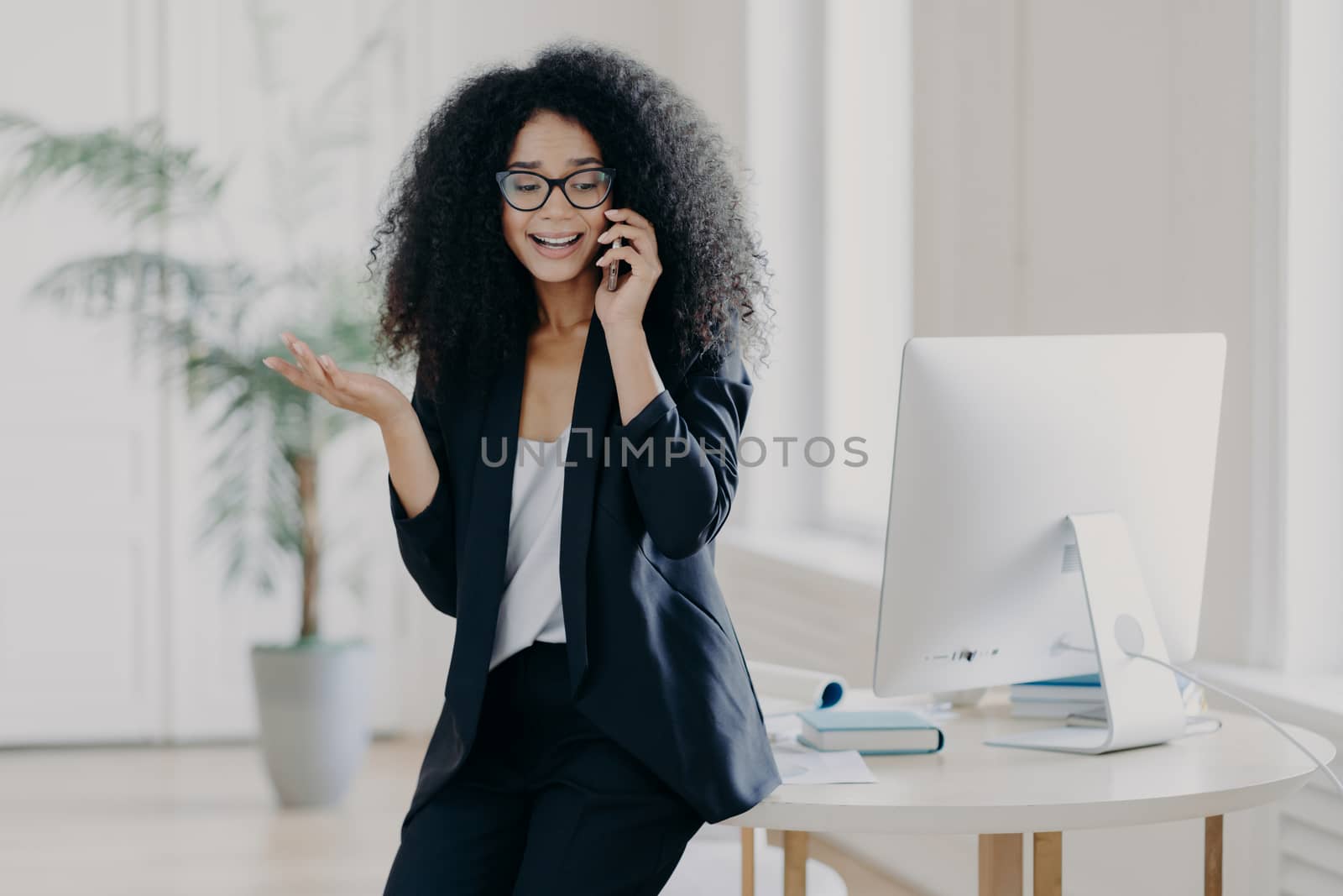 Photo of puzzled prosperous businesswoman calls partner, raises palm, holds mobile phone, wears spectacles and formal black outfit, tries to solve problem at work, discusses something actively by vkstock