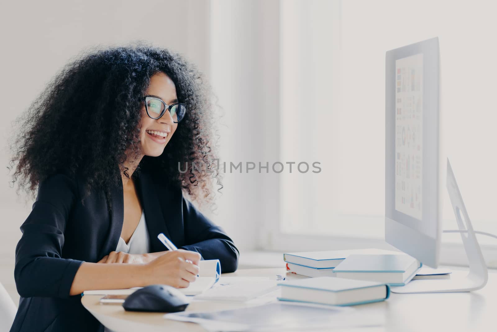 Professional curly haired woman manager makes report, focused into screen, writes down information, wears glasses and formal suit, notes idea for strartup planning, poses in office interior.