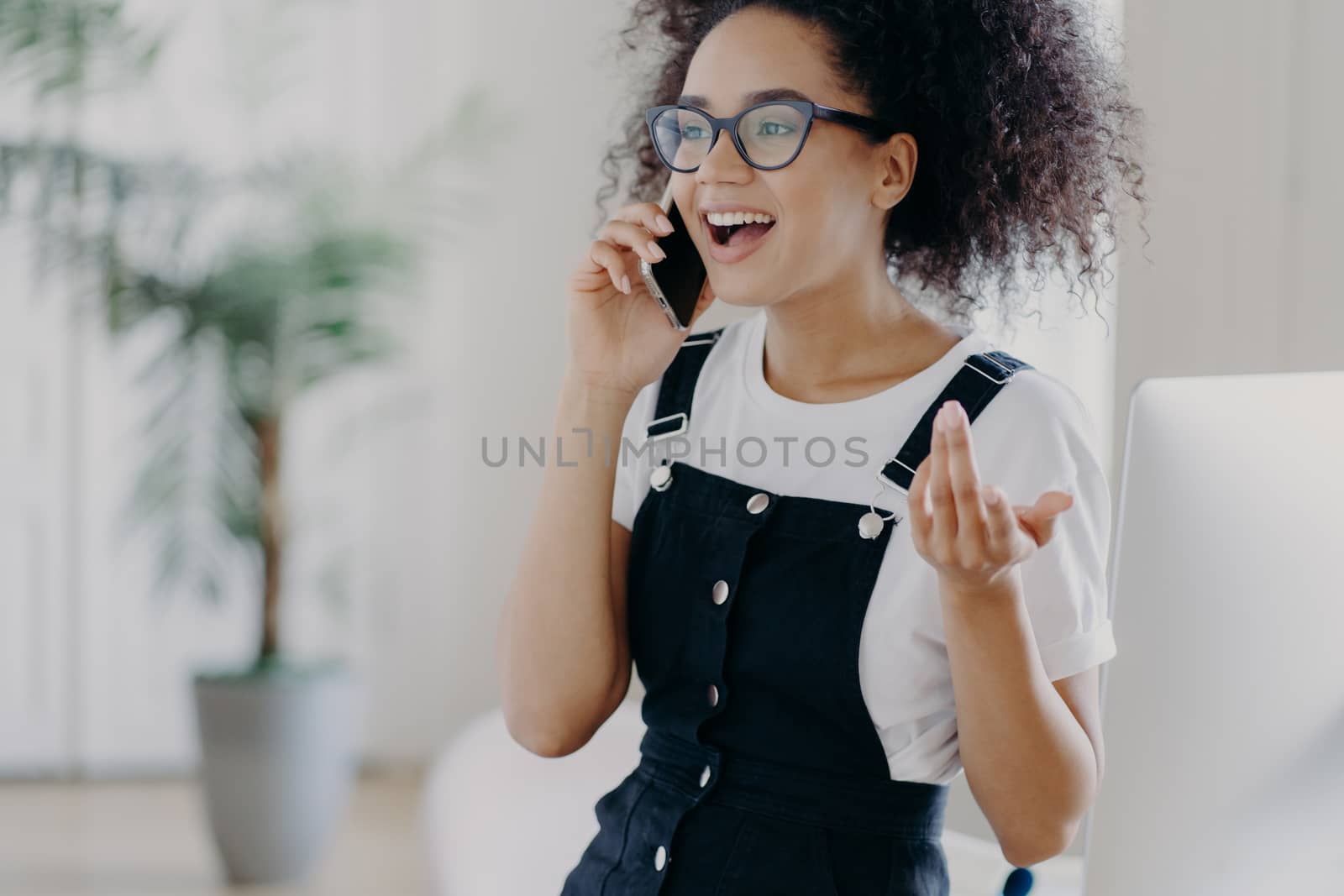 Cropped image of happy curly teenager has active discussion of something via cell phone, wears spectacles and sarafan, raises arm, gesticulates during phone conversation, has joyful expression by vkstock