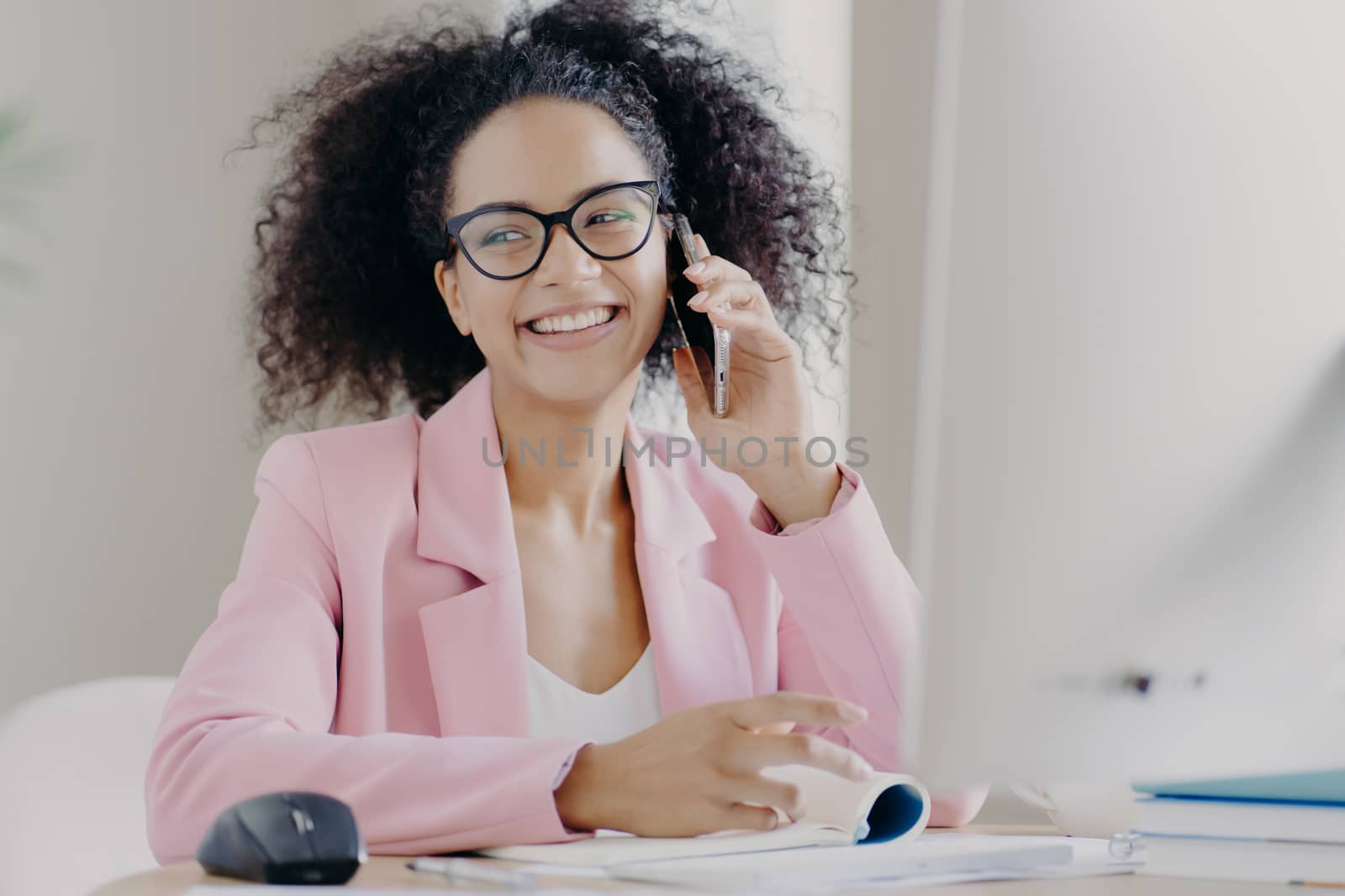 Pretty curly haired dark skinned woman enjoys telephone conversation with colleague, has happy expression, sits at desktop, wears glasses and rosy suit, has break after hard work. Technology by vkstock