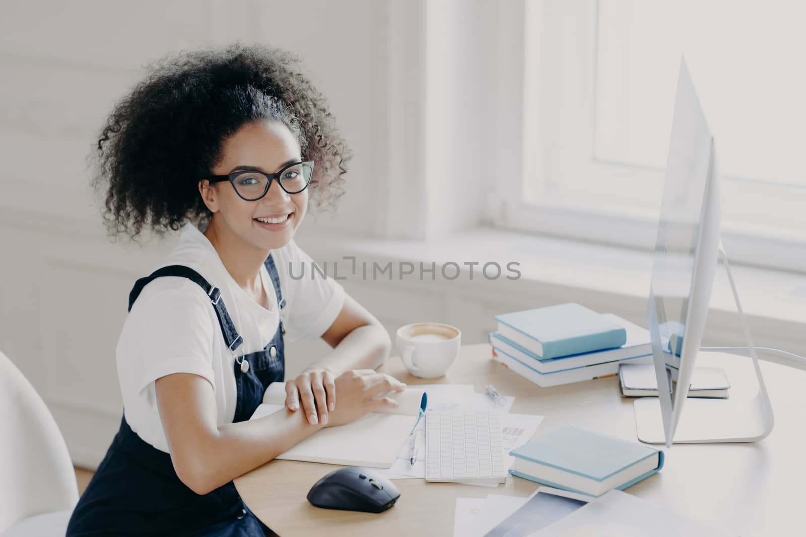 Positive Afro American female freelancer poses at workplace with papers and textbooks, works remotely on computer, has coffee break, works in her own cabinet, wears casual clothes. People, job by vkstock