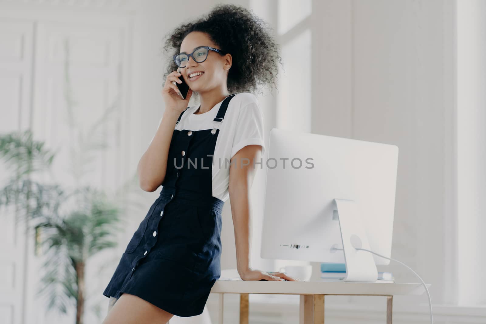 Pleased curly female office worker dressed in black sarafan, leans at table, poses in cabinet near computer monitor, has telephone conversation, discusses details of contract, has smile on face