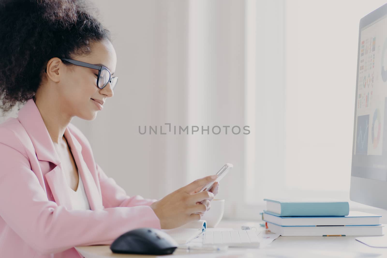 Horizontal shot of focused curly haired female office worker holds mobile phone, searches information in internet, wears optical glasses and elegant suit, computer monitor in front, does paper work by vkstock