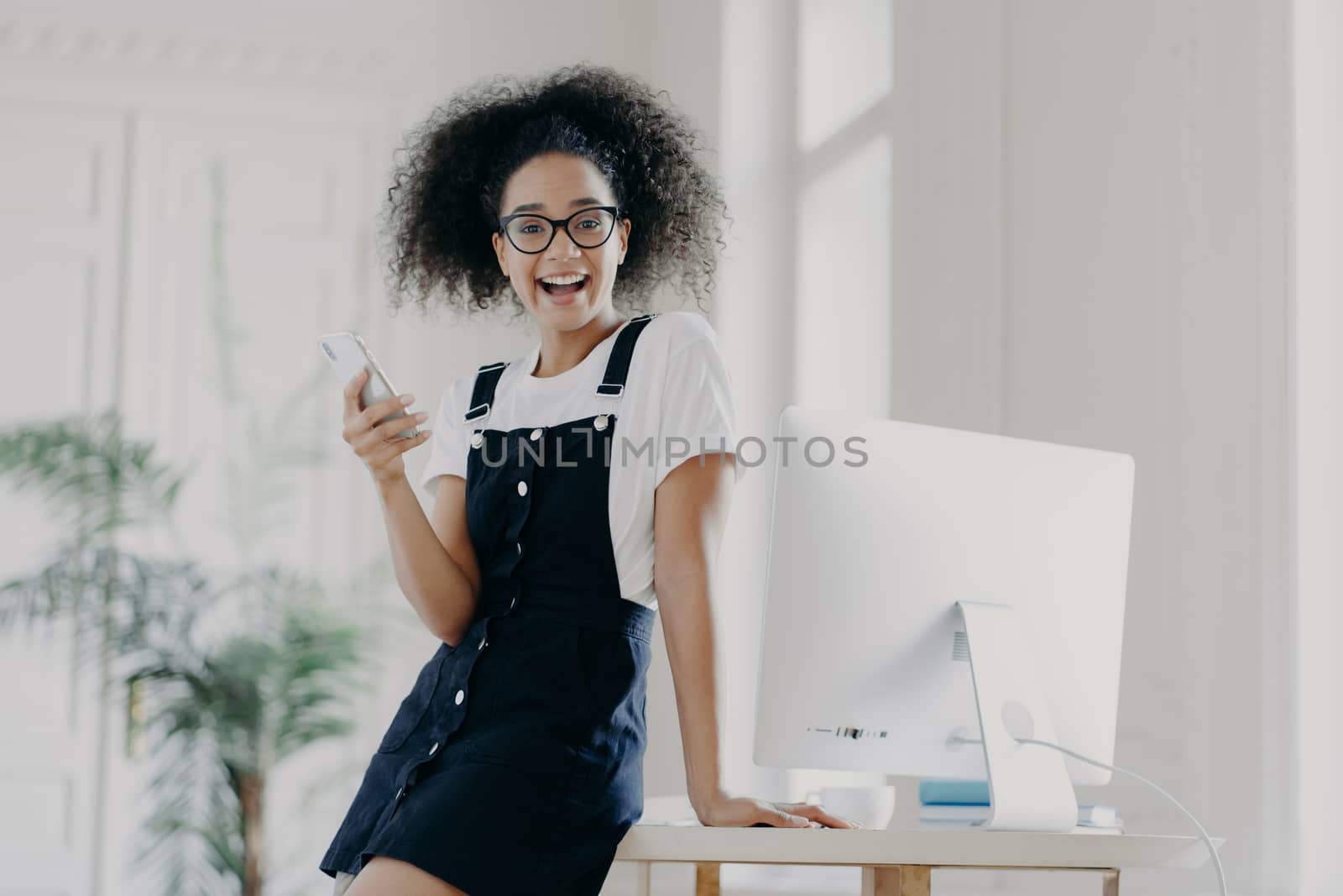 Happy carefree female financier uses mobile phone, glad to get salary in time, poses near wooden table, uses computer for work, wears white t shirt and sarafan, texts message, smiles pleasantly by vkstock