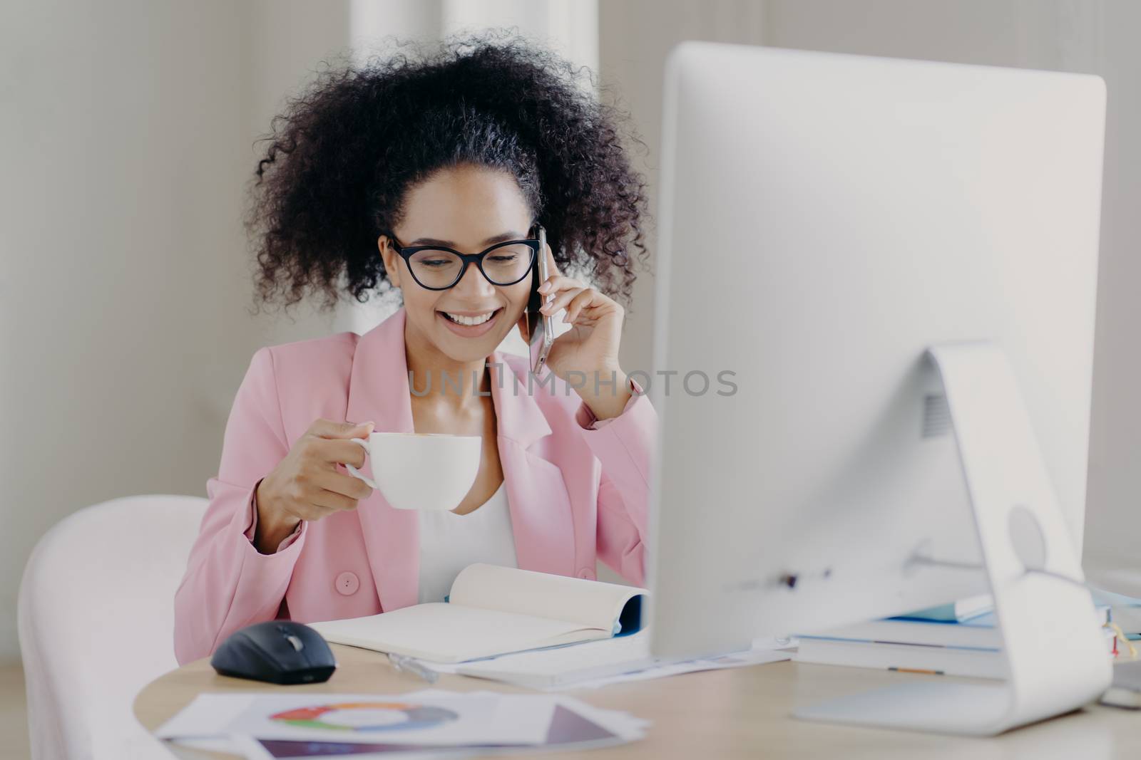 Happy dark skinned young woman with curly hair, makes telephone call, wears transparent glasses, dressed elegantly, holds mug of coffee, sits at workplace, poses near computer, poses in cabinet by vkstock