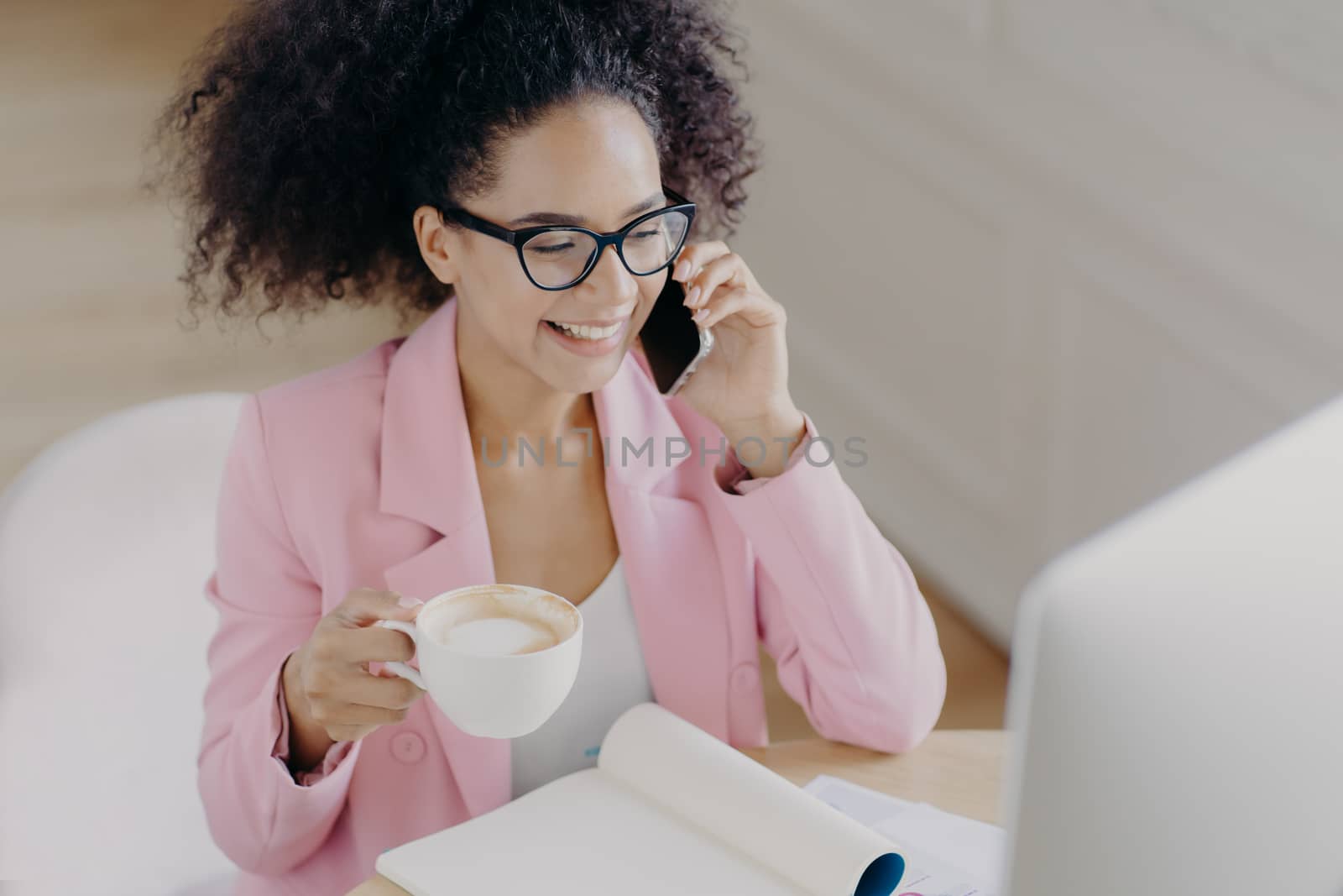 Top view of glad dark skinned woman with curly dark hair, has phone conversation, holds mug of drink, smiles pleasantly while looks at screen of computer, wears elegant attire, busy working at project by vkstock