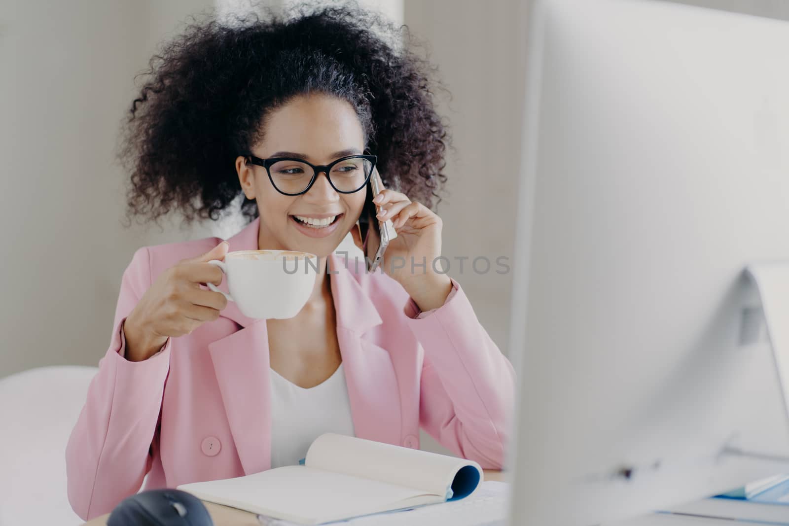 Photo of attractive smiling female entrepreneur enjoys aromatic beverage, holds white mug of drink, has telephone conversation, dressed in formal outfit, poses at workplace, wears transparent glasses by vkstock