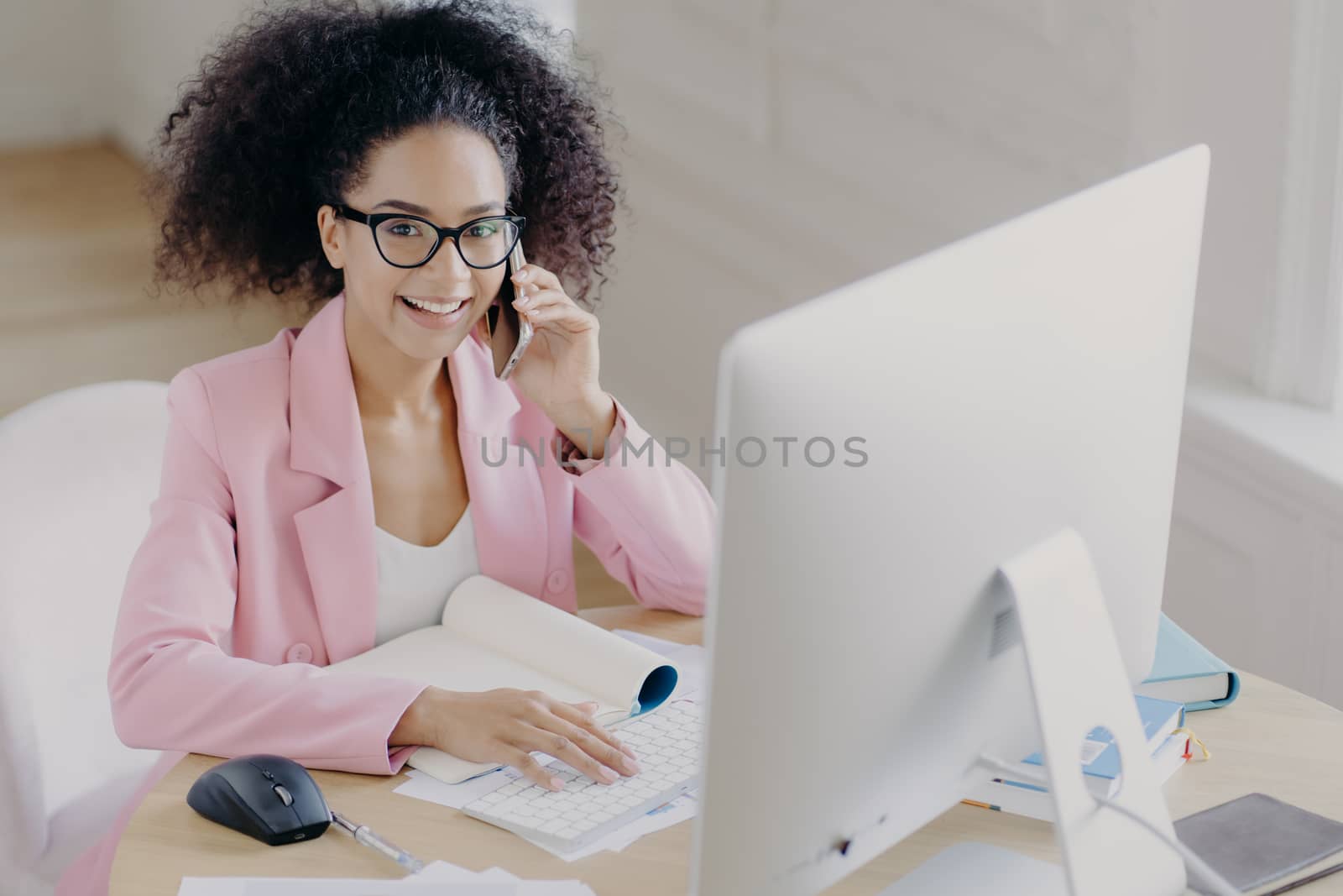 Positive dark skinned woman with curly combed hair, keybaords on computer, involved in working process, wears spectacles and elegant clothing, has telephone conversation, poses at workplace. by vkstock