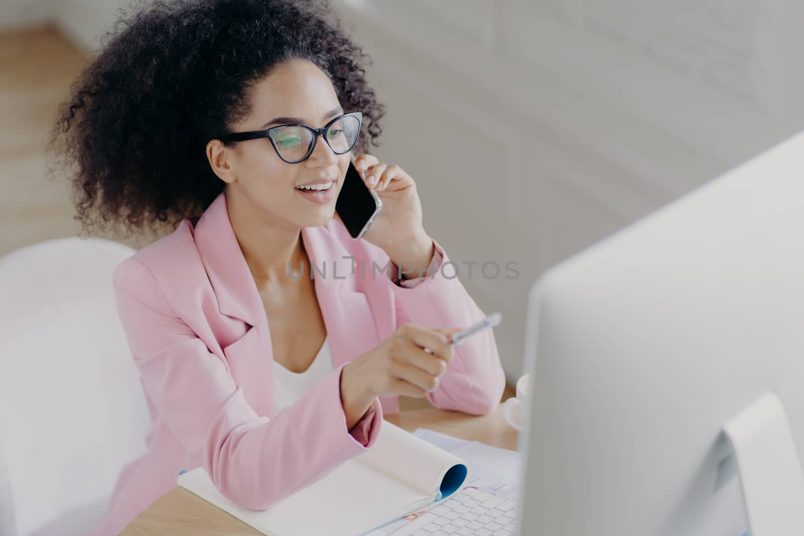 Glad young African American woman discusses business news, points with pen at screen on computer, talks via modern cell phone, busy with paper work, wears transparent glasses and formal wear