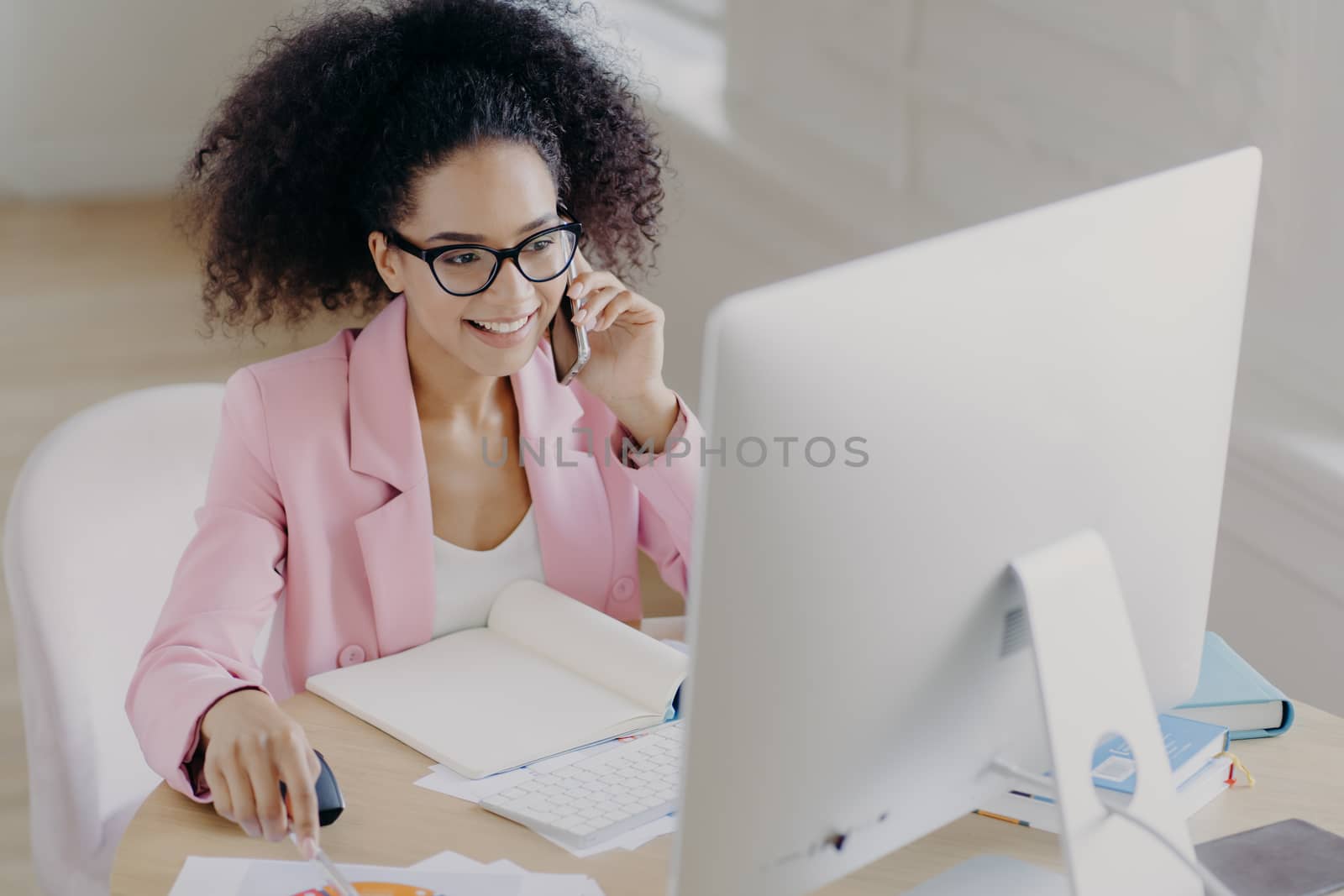 Photo of happy curly haired female wears optical glasses, pink jacket, looks attentively at computer display, sits at desktop with opened notepad, calls someone via smartphone, has positive smile by vkstock