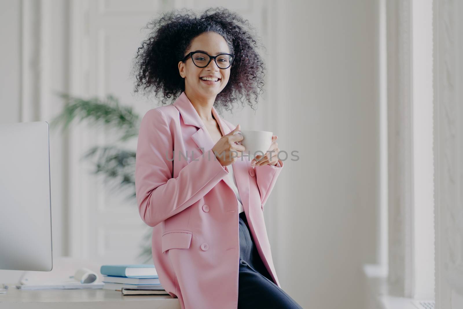 Photo of relaxed businesswoman holds cup of hot drink, has coffee break, stands near her workplace in spacious white cabinet wears spectacles long pink jacket works in office. Time for rest after work by vkstock