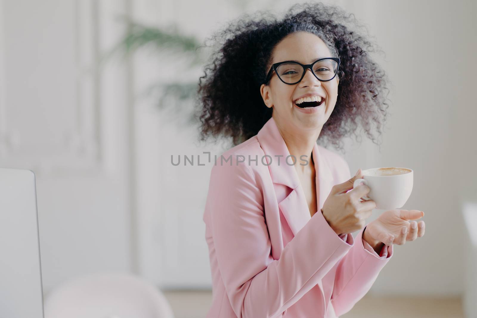 Overjoyed curly haired woman laughs happily while drinks hot coffee or latte, wears transparent spectacles, has fun during break in office, shows white teeth, being professional entrepreneur by vkstock