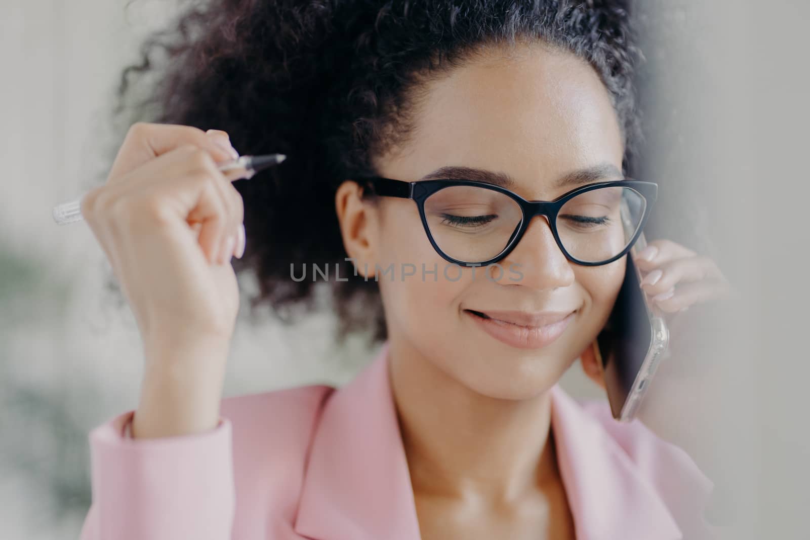 Headshot of delighted female director holds pen in hand, wears optical eyeglasses, has gentle smile, makes call via modern cell phone, dressed in pink elegant costume, focused down with shyness by vkstock