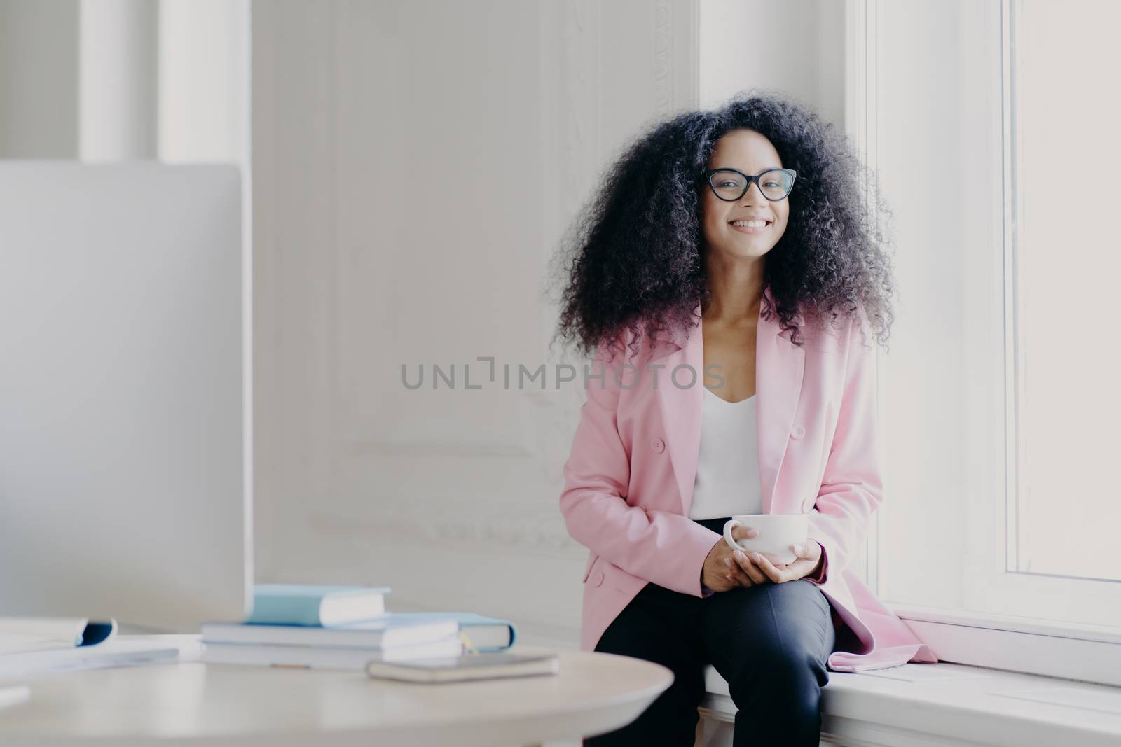 Happy female entrepreneur has bushy Afro hair, wears rosy jacket and black trousers, holds cup of drink, poses at windowsill in spacious cabinet with table, modern computer, enjoys aromatic coffee by vkstock
