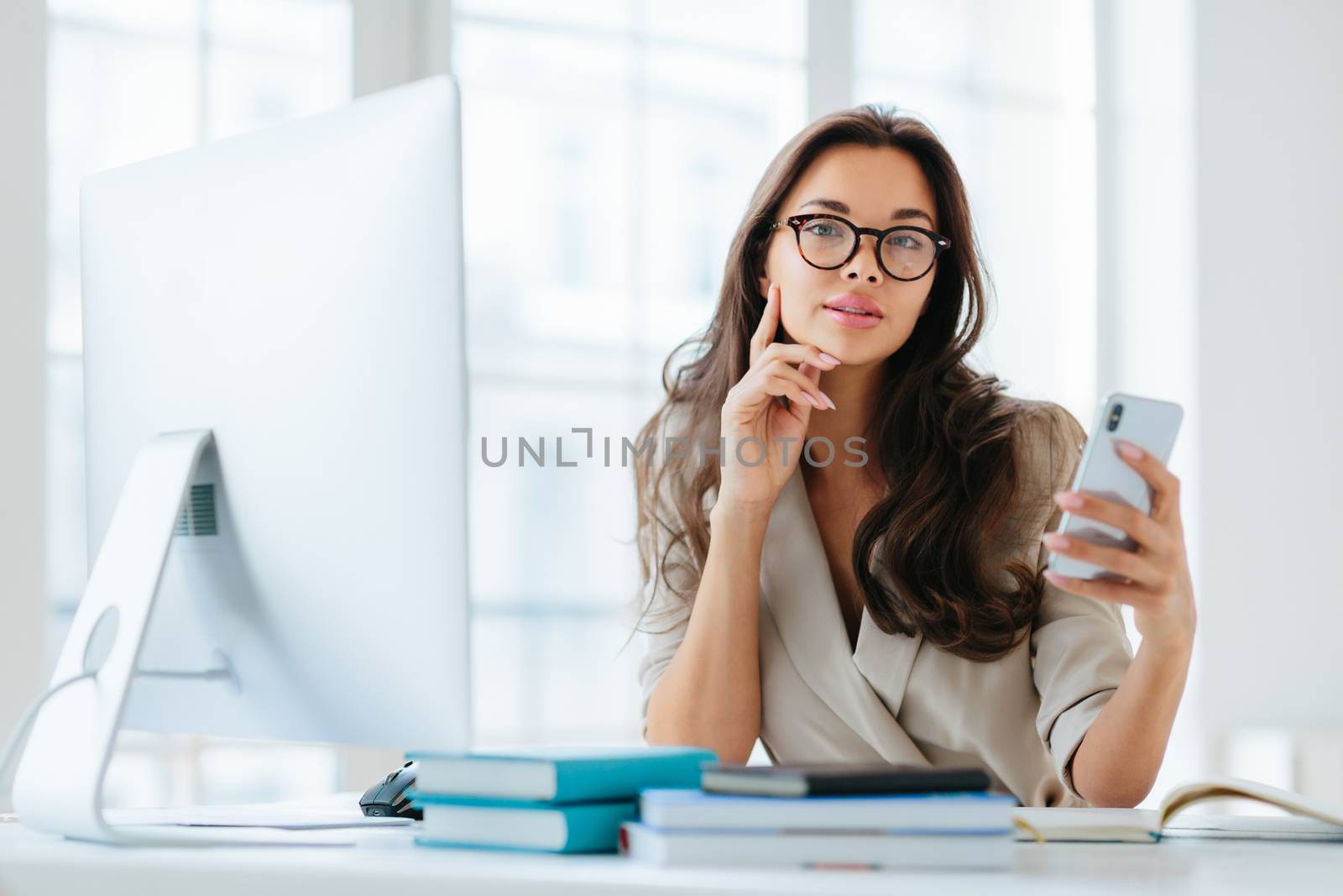 Female company office worker in formal wear uses modern gadget for work, poses at workplace near monitor, plans working schedule, browses information online, poses against window. Working time by vkstock