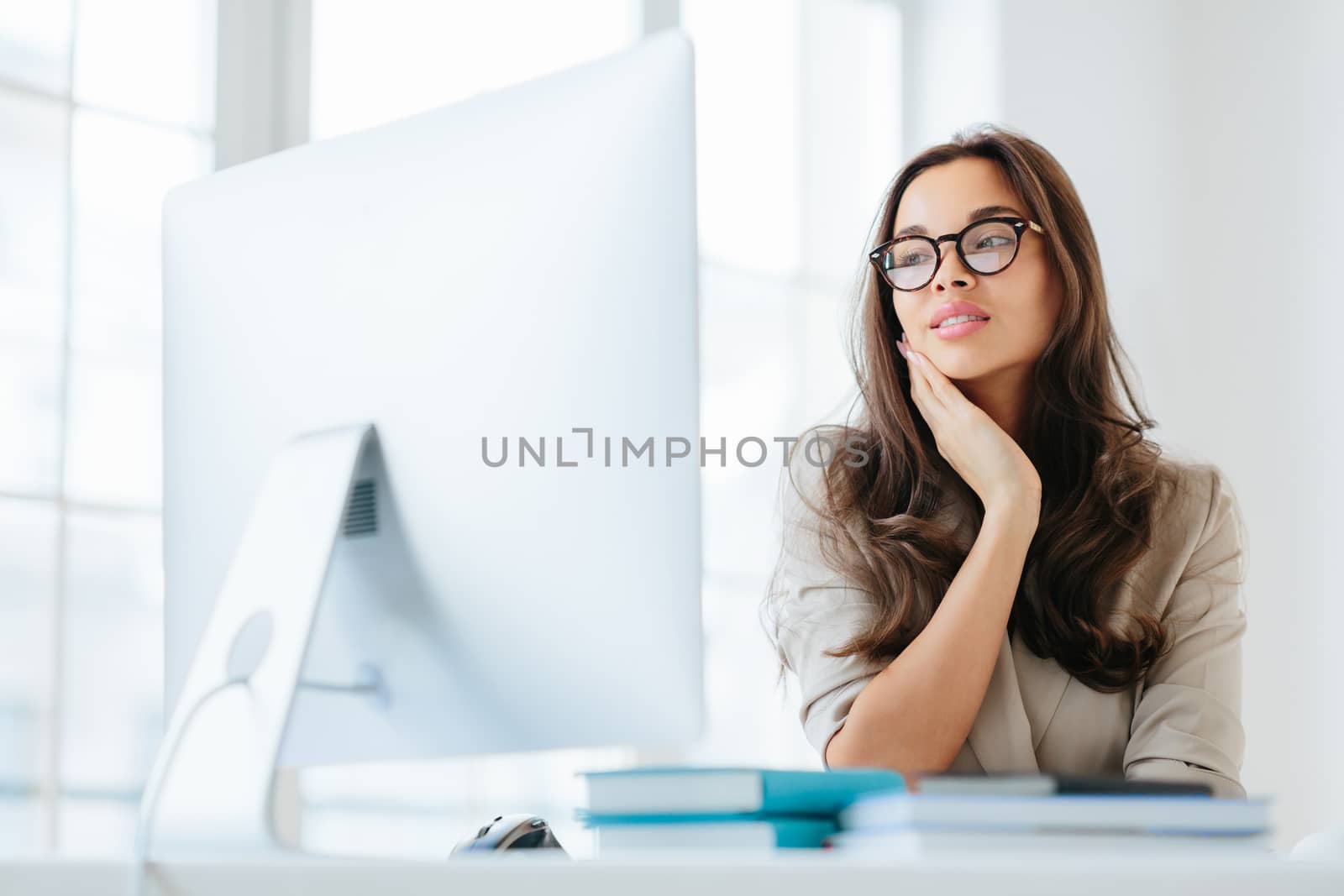 Indoor shot of serious brunette lady focused in monitor of computer, uses wireless internet for publication, keeps hand on cheek, has attentive look, sits at desktop, works on business contract by vkstock