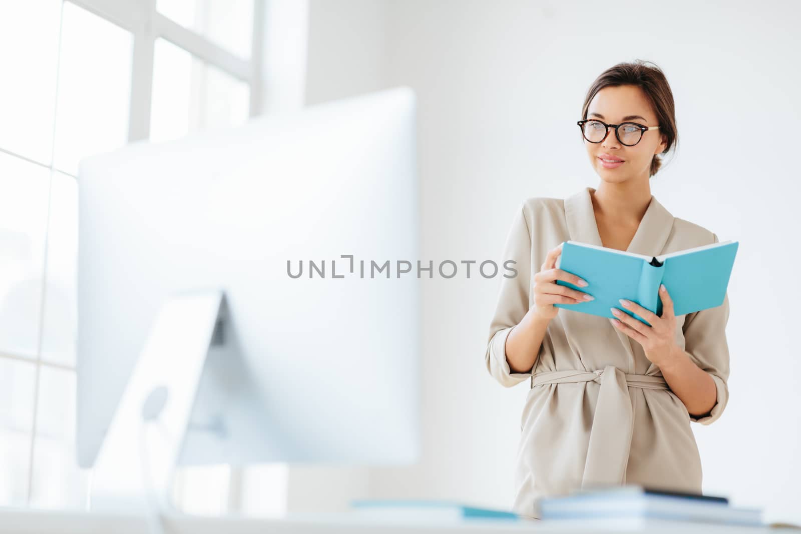 Young dark haired woman in elegant wear holds blue notepad focused into monitor, ponders about business strategy for eneterprise, wears transparent eyeglasses, poses in spacious white own cabinet by vkstock