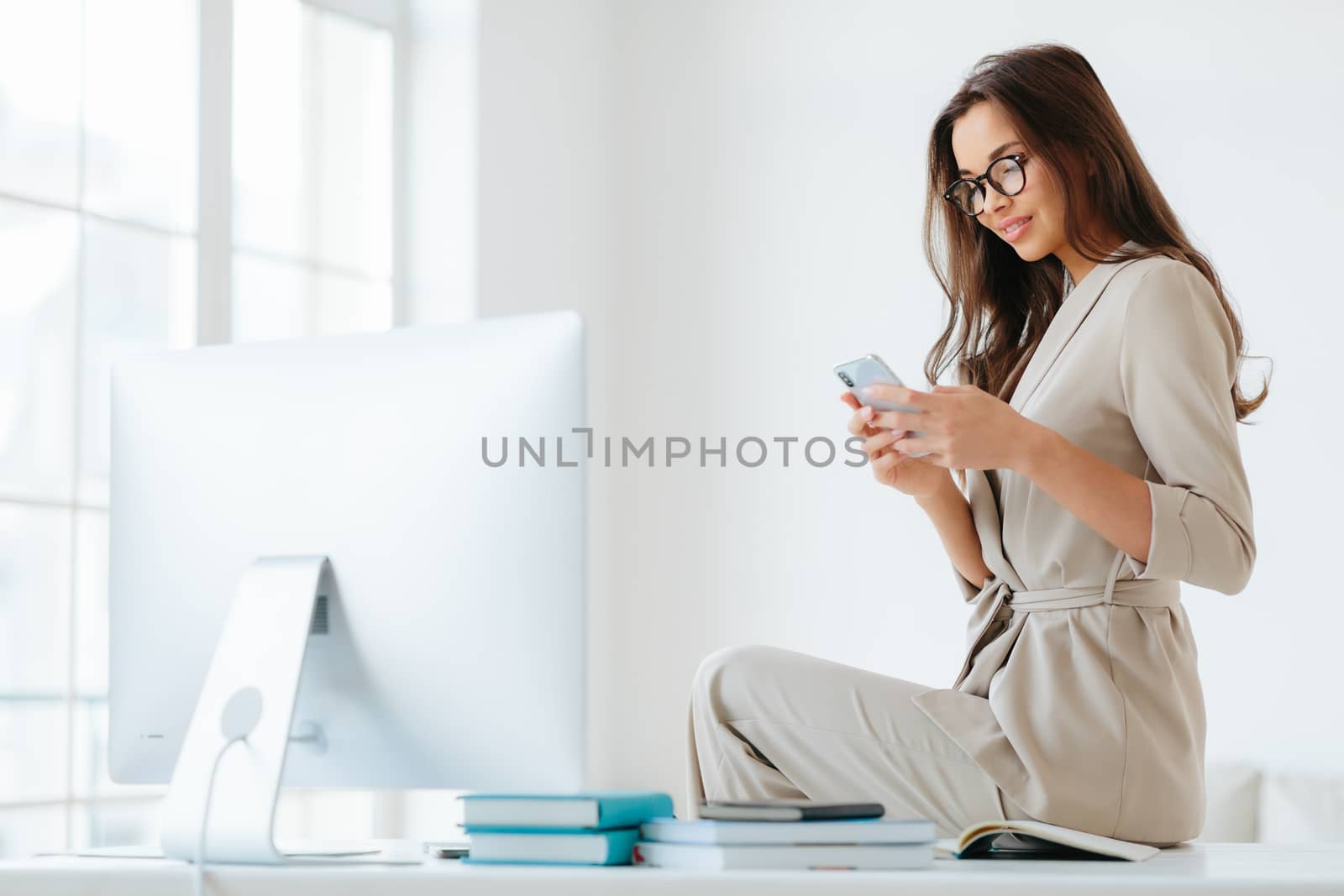 Horizontal shot of beautiful young woman in elegant clothes, has gentle smile, checks newsfeed via modern smartphone, has rest after hard work, poses in office interior at desktop, white walls