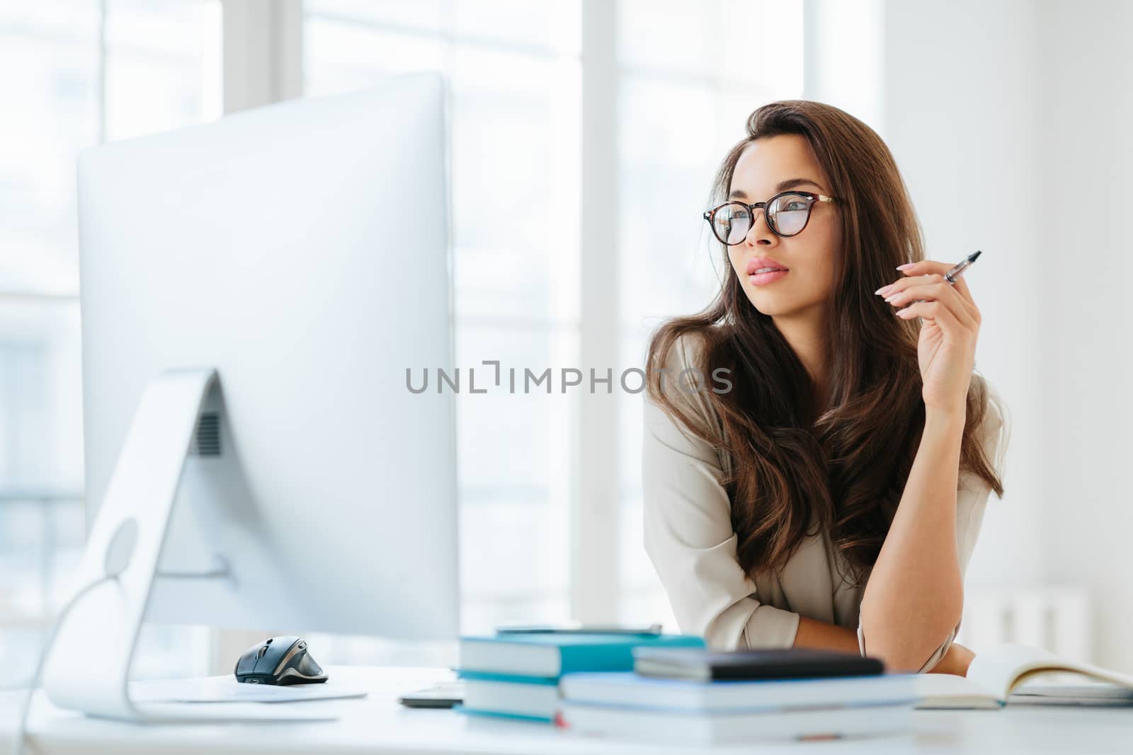 Dark haired lady focused into big monitor, sits at desktop, holds pen and writes notes, wears spectacles for vision correction, poses in coworking space. Femlae manager at workplace, makes research