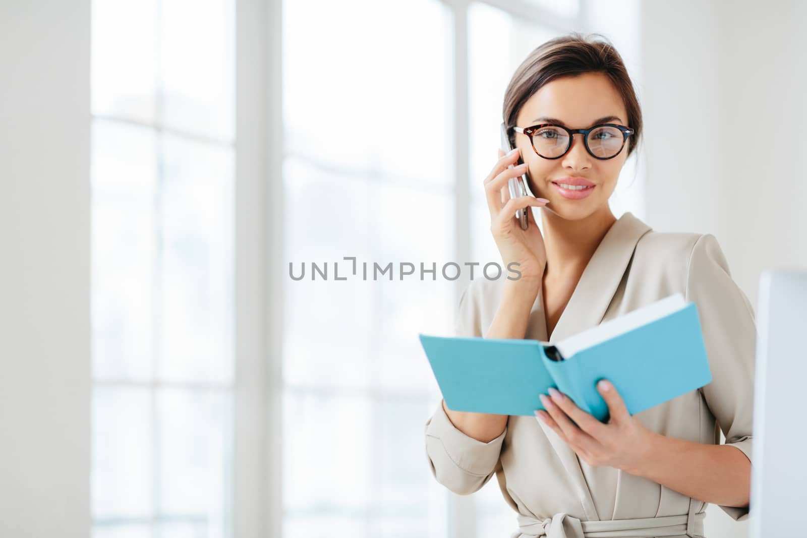 Photo of attractive woman with dark hair, has telephone conversation, agrees to meet with colleague, holds opened diary, wears glasses and formal suit, poses over white background plans daily schedule