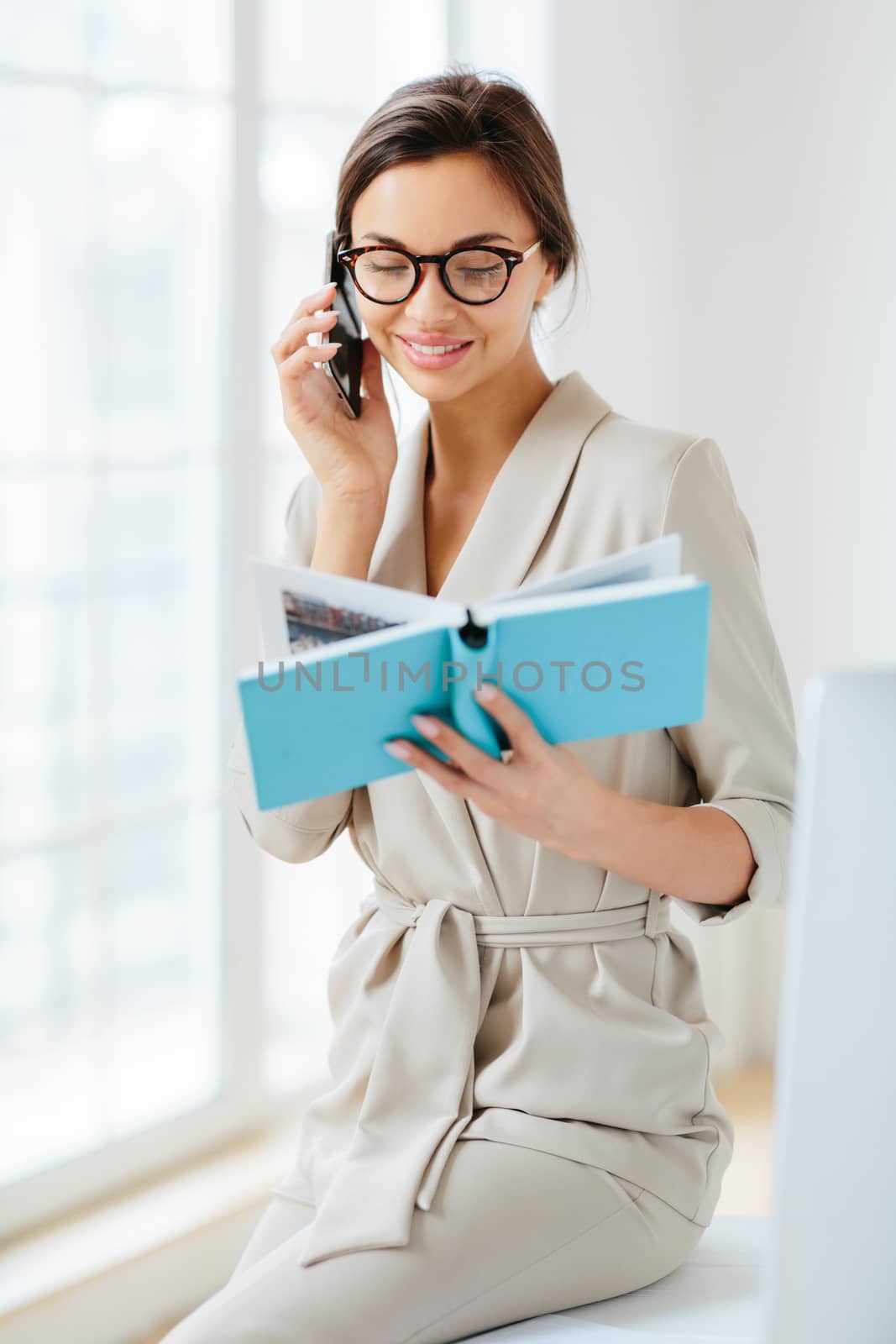 Vertical shot of female administrative manager involved in working process, holds notepad with written notes, discusses something via cellphone, wears spectacles and elegant costume, smiles gently by vkstock