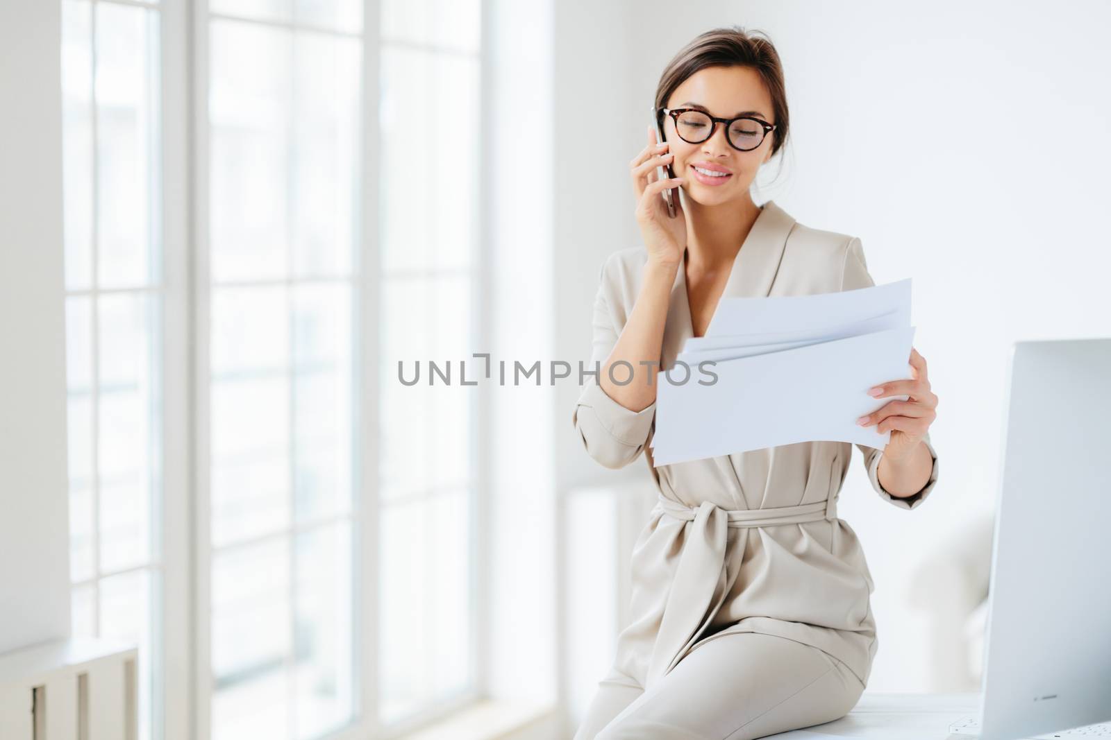 Attractive businesswoman reads papers or business documents, has telephone talk with business partner, examines paperwork before meeting, sits at desktop, smiles happily, works with documentation