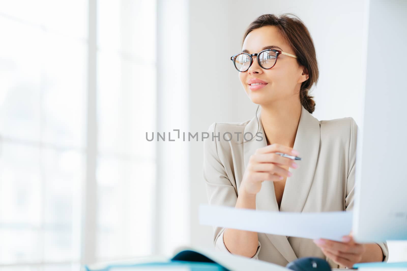 Photo of thoughtful brunette woman in formal wear, spectacles, holds pen and paper, looks pensively away, thinks about development of own business, works freelance, poses in coworking space. by vkstock