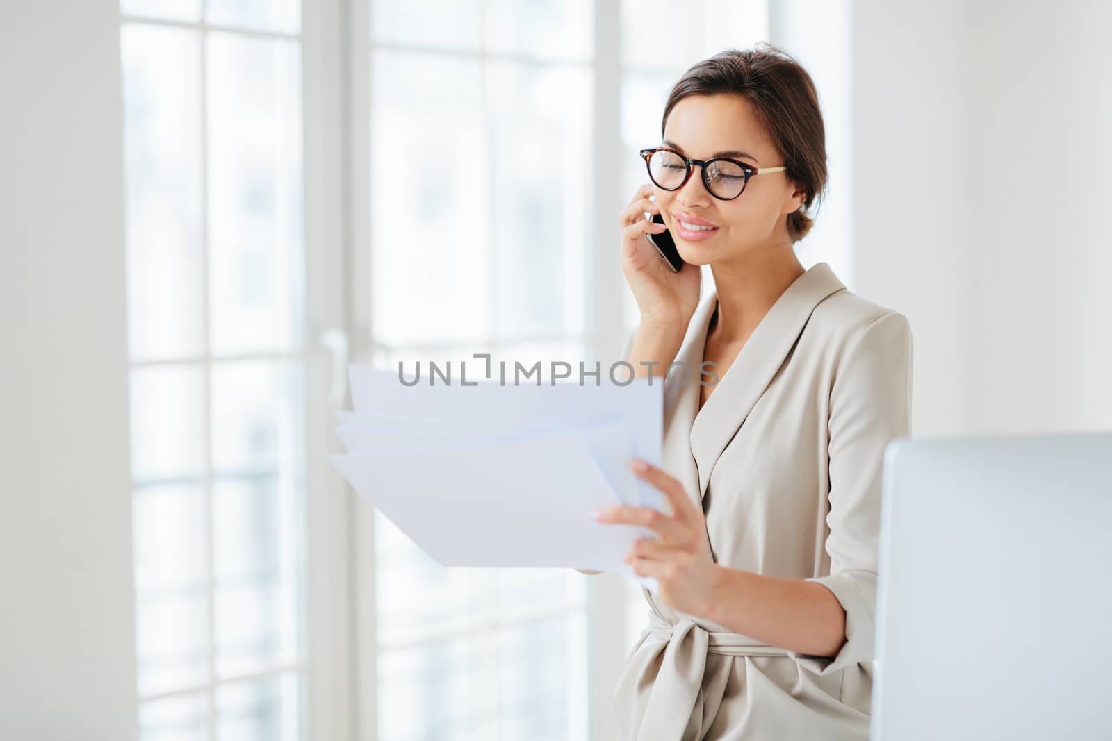 Positive woman in formal clothes, calls to business partner, discusses financial startup project, reads information from papers, wears optical glasses, stands in office, works at international company by vkstock