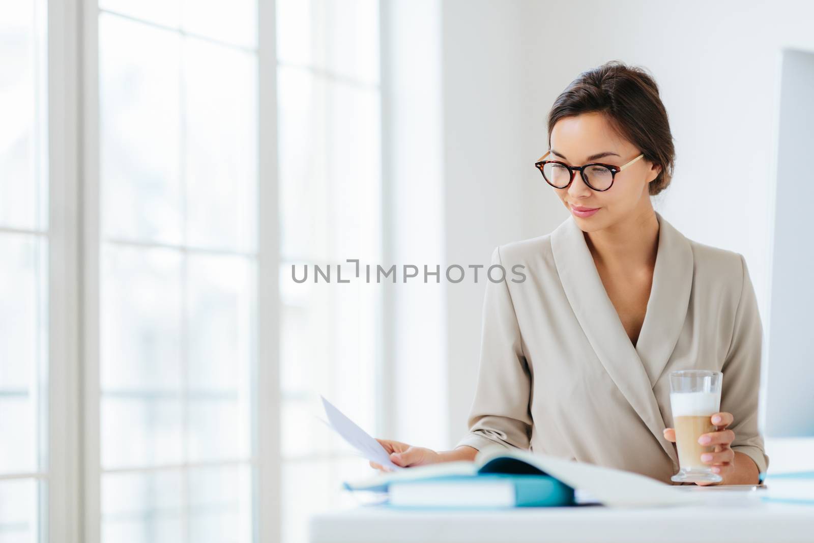 Woman economist checks information on business paper, wears spectacles and elegant suit, drinks milkshake, poses against office interior, being in cabinet, prepares startup project. Working process