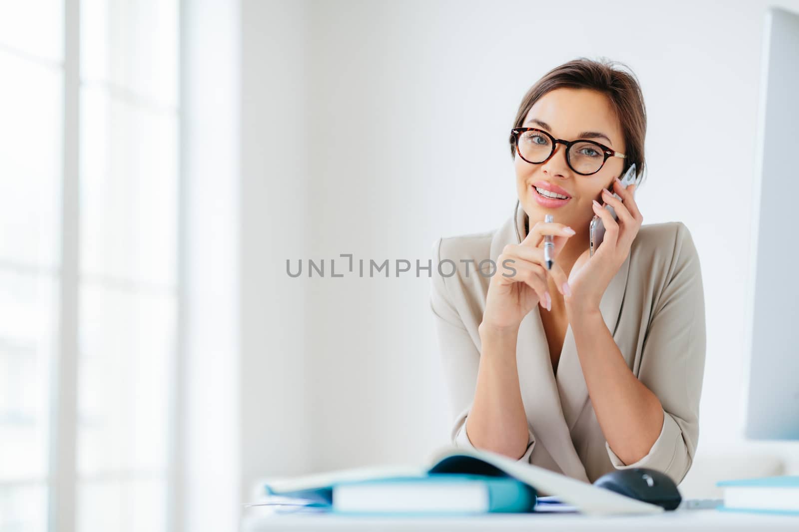 Photo of female employee has cheerful expression, talks via smartphone, hold pen, sits at desktop with papers and notepads, computer, poses against office interior, wears spectacles, elegant clothes