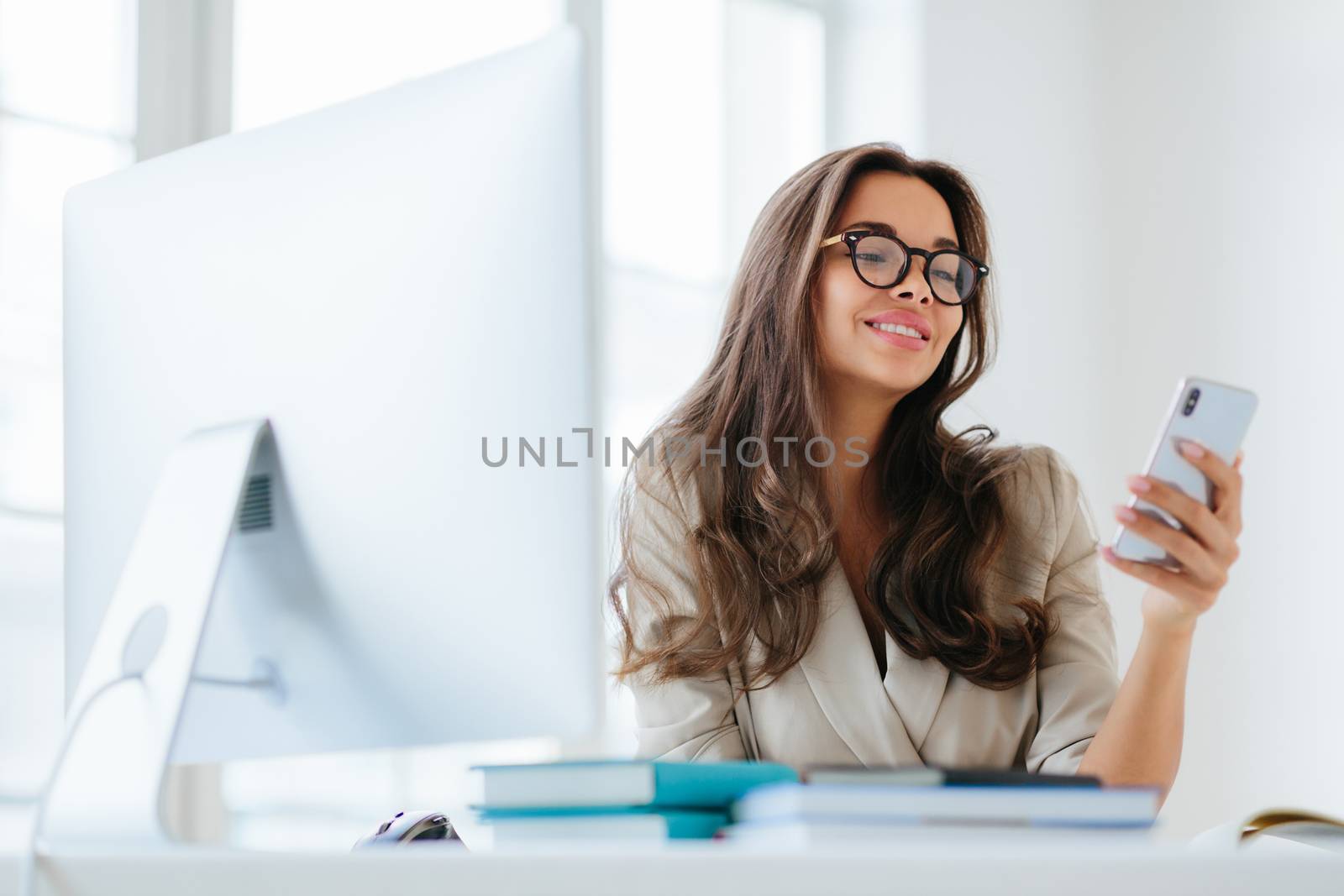 Smiling female in corporate clothes watches funny video via smartphone, sits at desktop in office, uses computer for work, enjoys working process, browses information for project, wears eyeglasses by vkstock