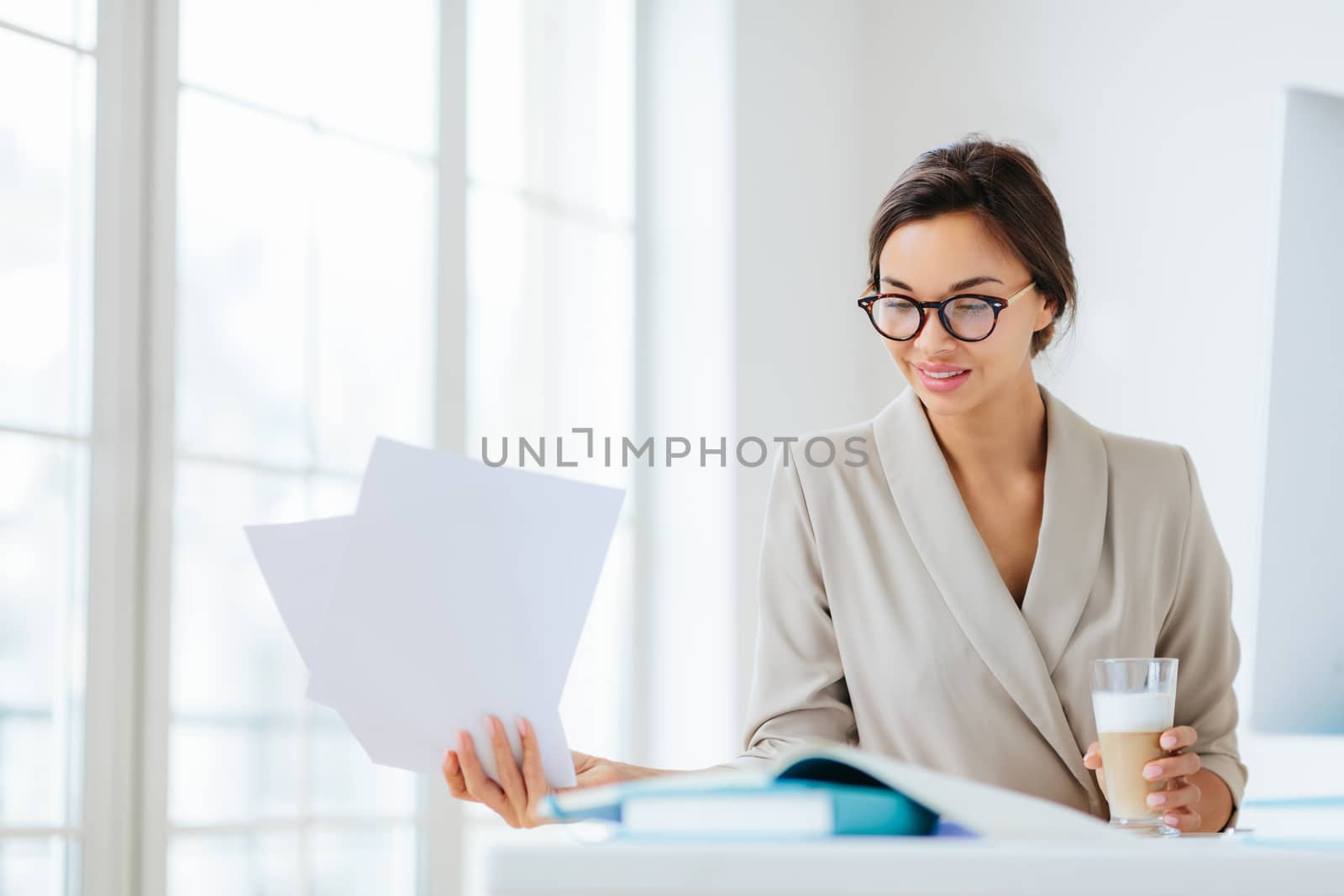 Horizontal view of confident brunette female worker in formal wear and optical spectacles looks through documentation, drink fresh beverage, focused down, poses indoor, has pleased expression