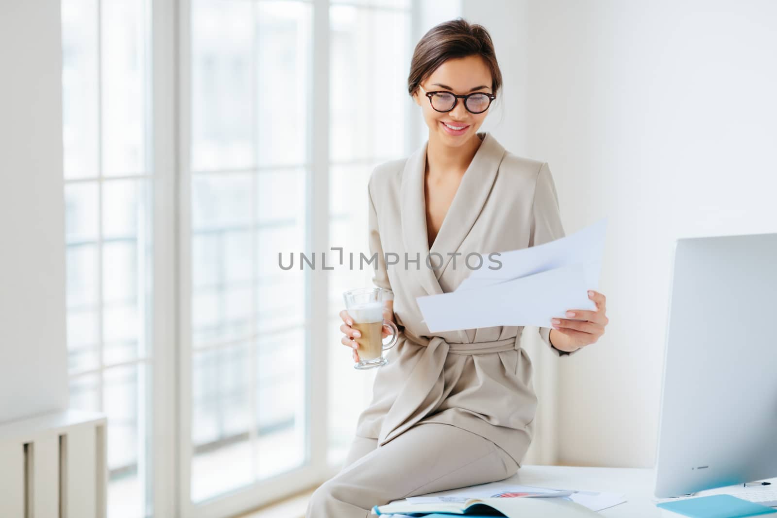 Pleased dark haired woman with smile, checks bills in documents, dressed beige business suit, drinks beverage, sits at desktop near big monitor, holds paper sheet with charts, poses near window