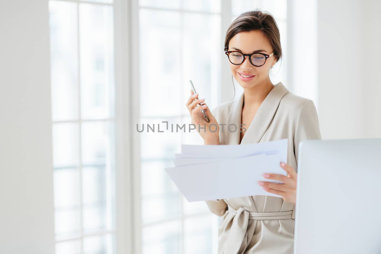 Photo of satisfied young woman with dark hair dressed in business suit, focused in papers, works in office, holds modern cellphone, wears optical glasses for good vision, has pleased expression