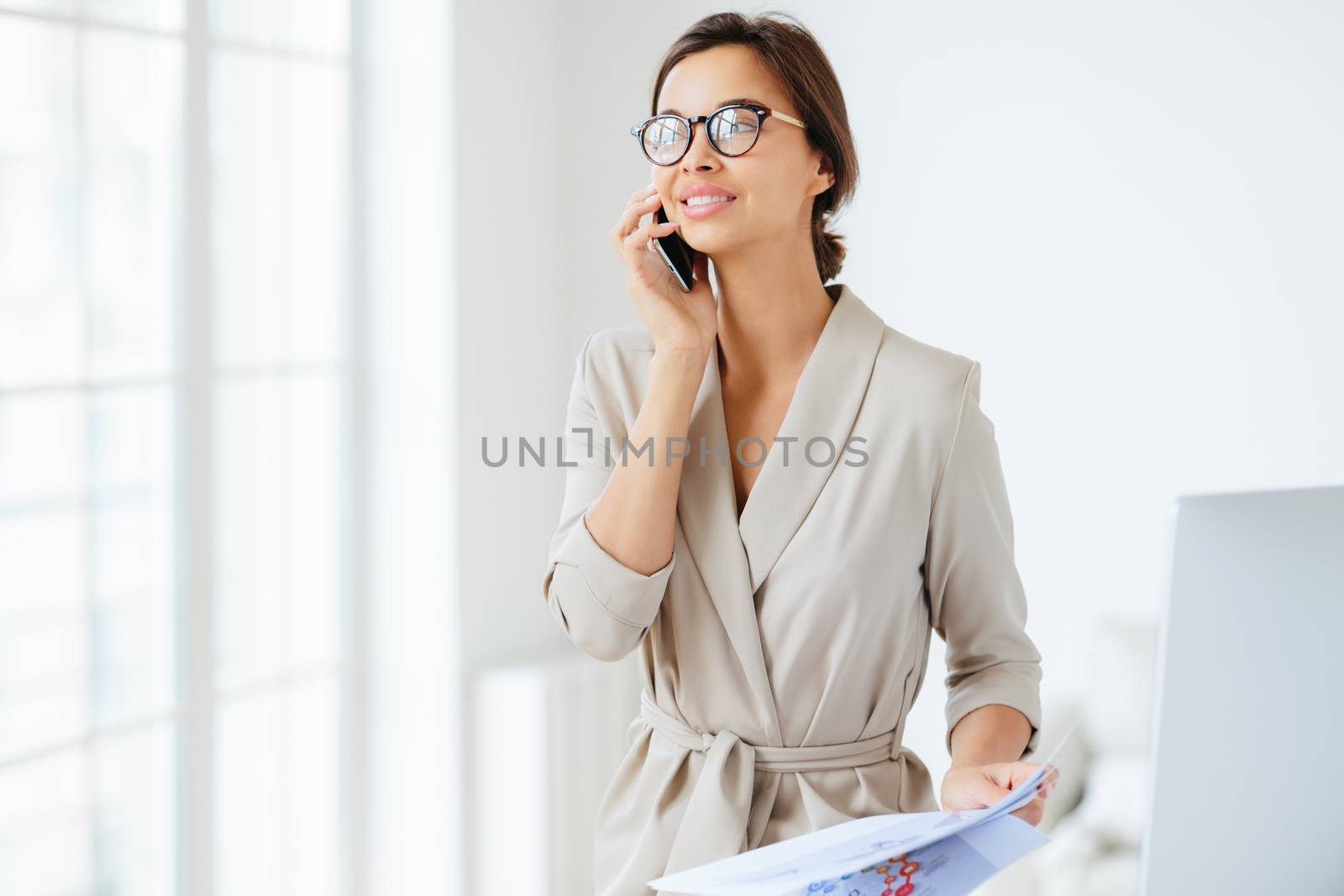Happy thoughtful successful female employee holds modern cellphone near ear, has telephone conversation, looks aside, dressed elegantly, reviews monthly report, studies graphics, stands in office by vkstock