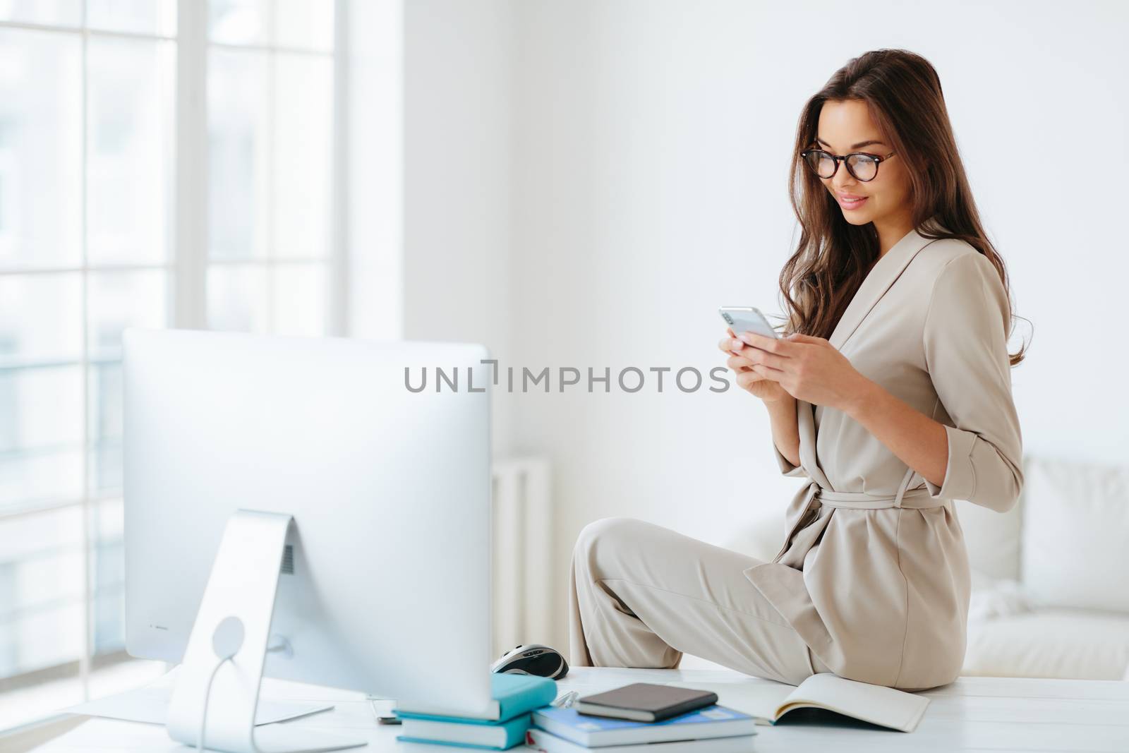 Elegant woman in beige formal suit reads news on website attentively, sits at desktop alone in cabinet, computer monitor and notepads around. Female employee uses modern smartphone in office by vkstock