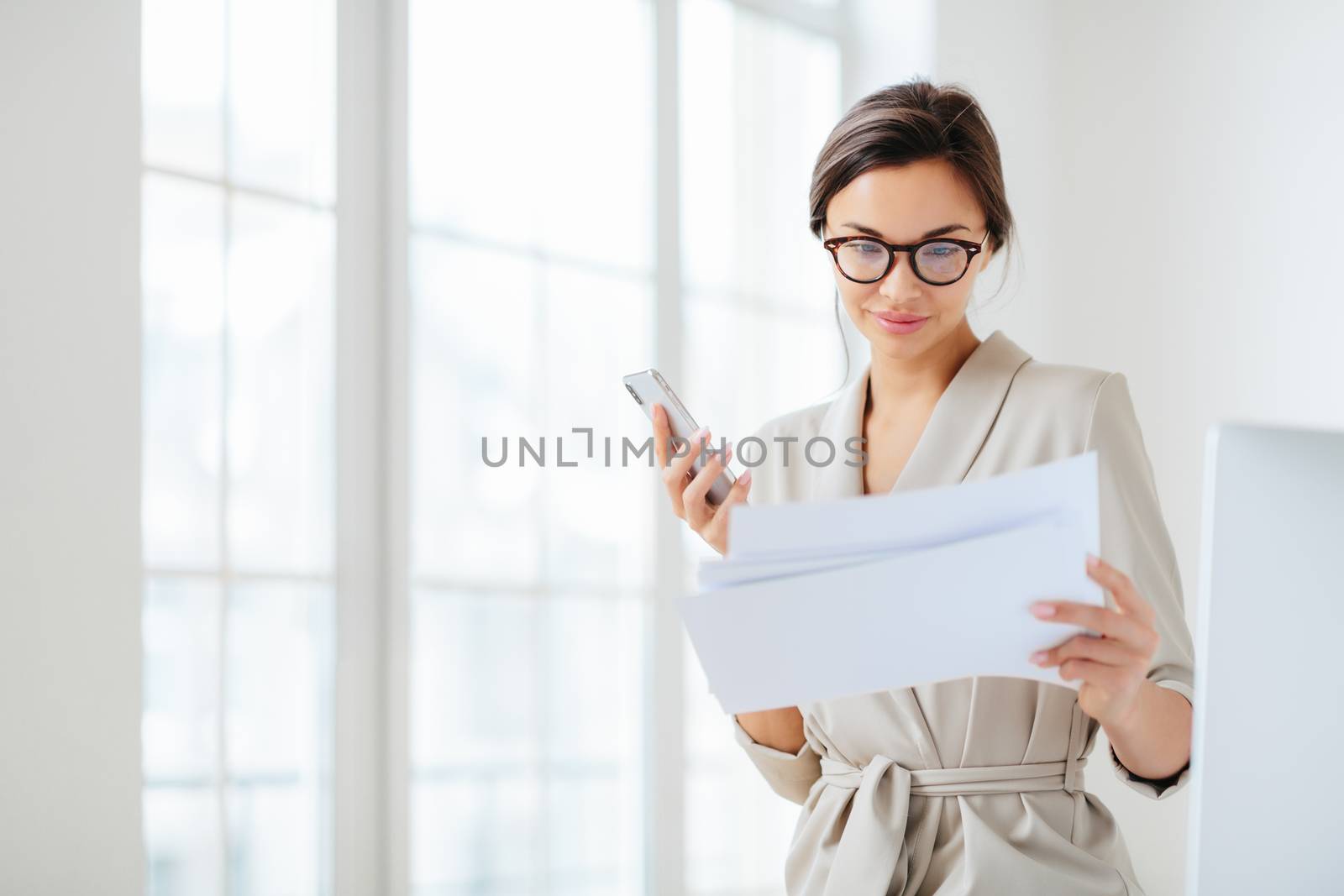 Satisfied female employee focused in documents, reads prepared report attentively, holds modern smartphone, wears transparent glasses and business suit poses over office interior checks monthly income