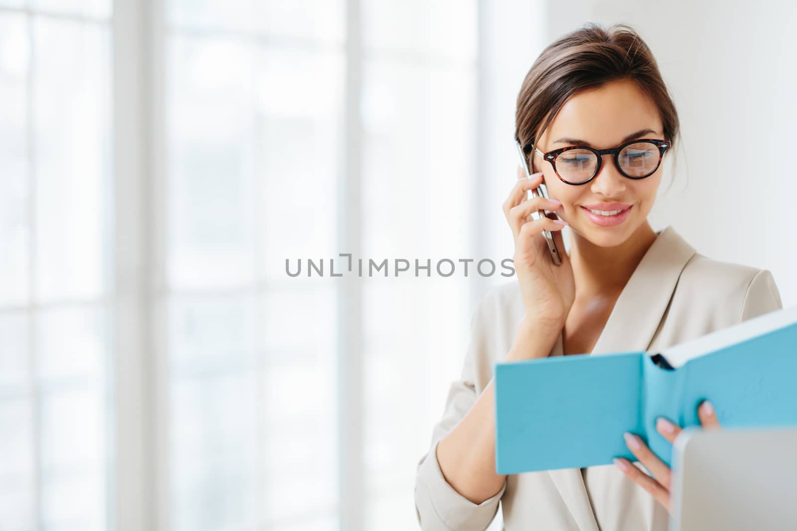 Photo of satisfied brunette woman in transparent glasses has telephone conversation, holds cellular, wears optical glasses, formal wear, poses against white blurred background, looks in notepad