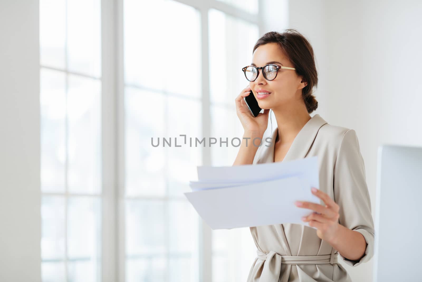 Pensive businesswoman stands sideways to camera, holds paper documents, has phone talk, prepares financial report, poses in coworking space, wears optical glasses and formal suit. Paperwork.