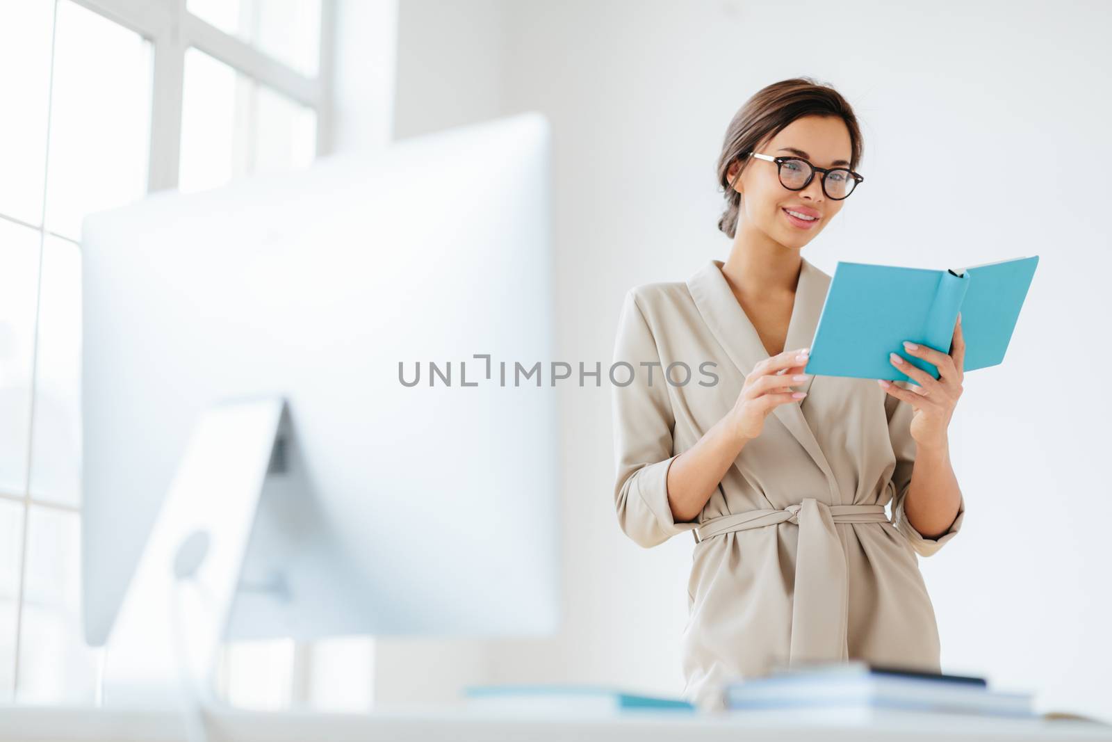 Cheerful female director holds personal organizer, looks through plans for day, wears beige formal costume, spectacles poses near dektop with big monitor white background verifies recorded information by vkstock