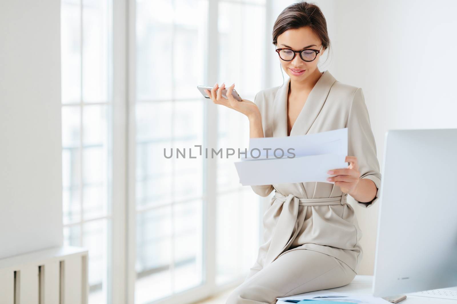 Photo of beautiful female entrepreneur in formal suit, focused in smartphone, checks information from paper documents, holds phone, going to search financial news online. Lawyer prepares for briefing
